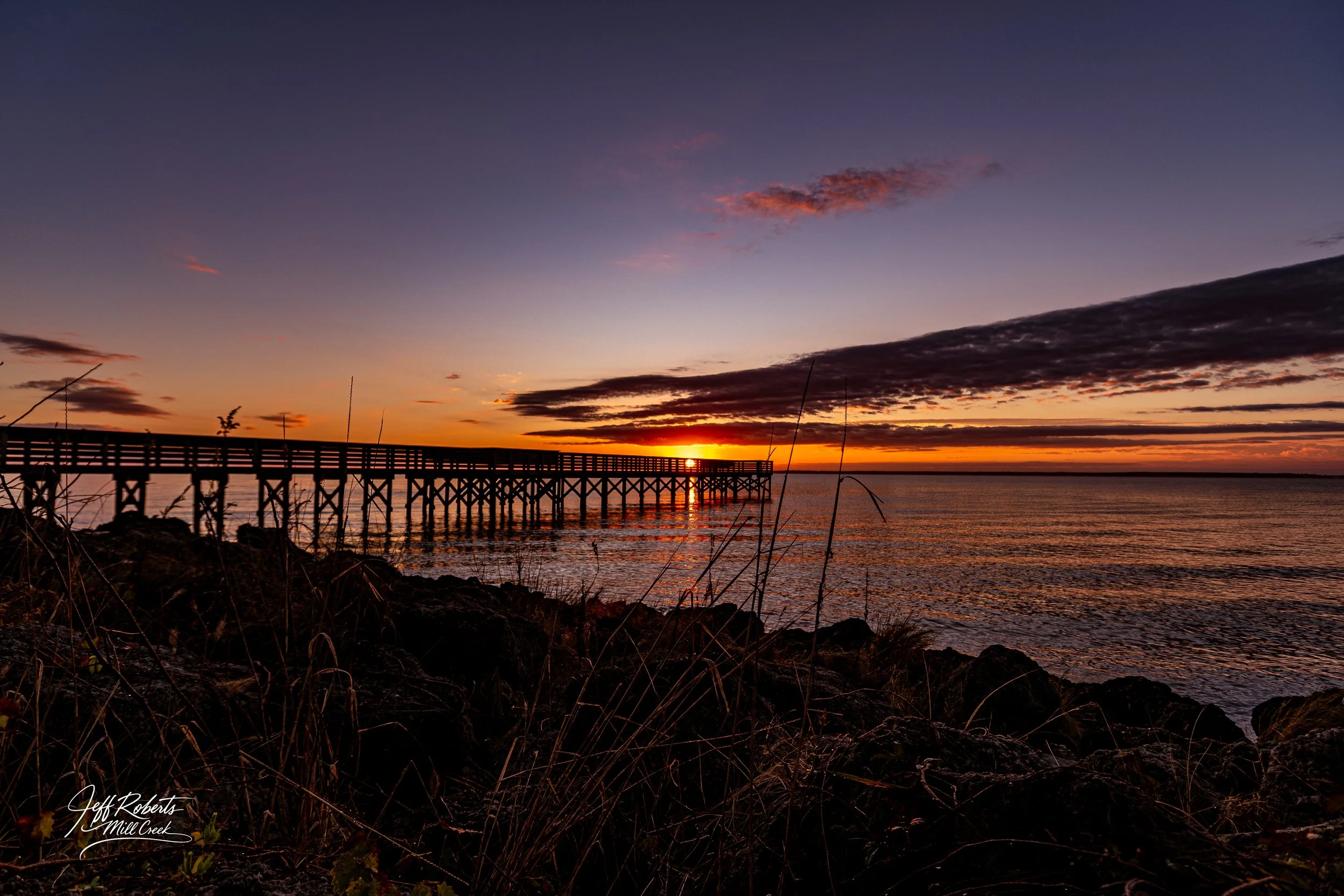 Sunset over a body of water with a wooden pier extending into the distance, dark clouds in the sky, and rocky shoreline with sparse vegetation in the foreground.