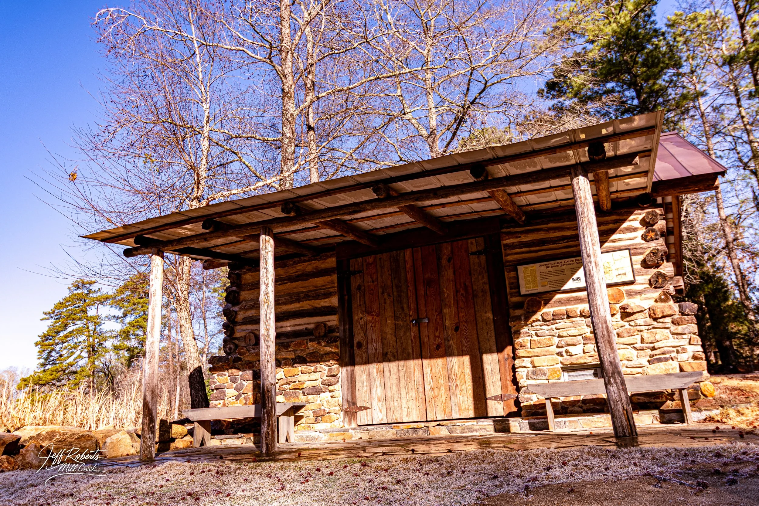 A small rustic log cabin with a wooden door, stone foundation, and a metal roof, situated in a wooded area with leafless trees and some evergreen trees in the background, under a clear blue sky.