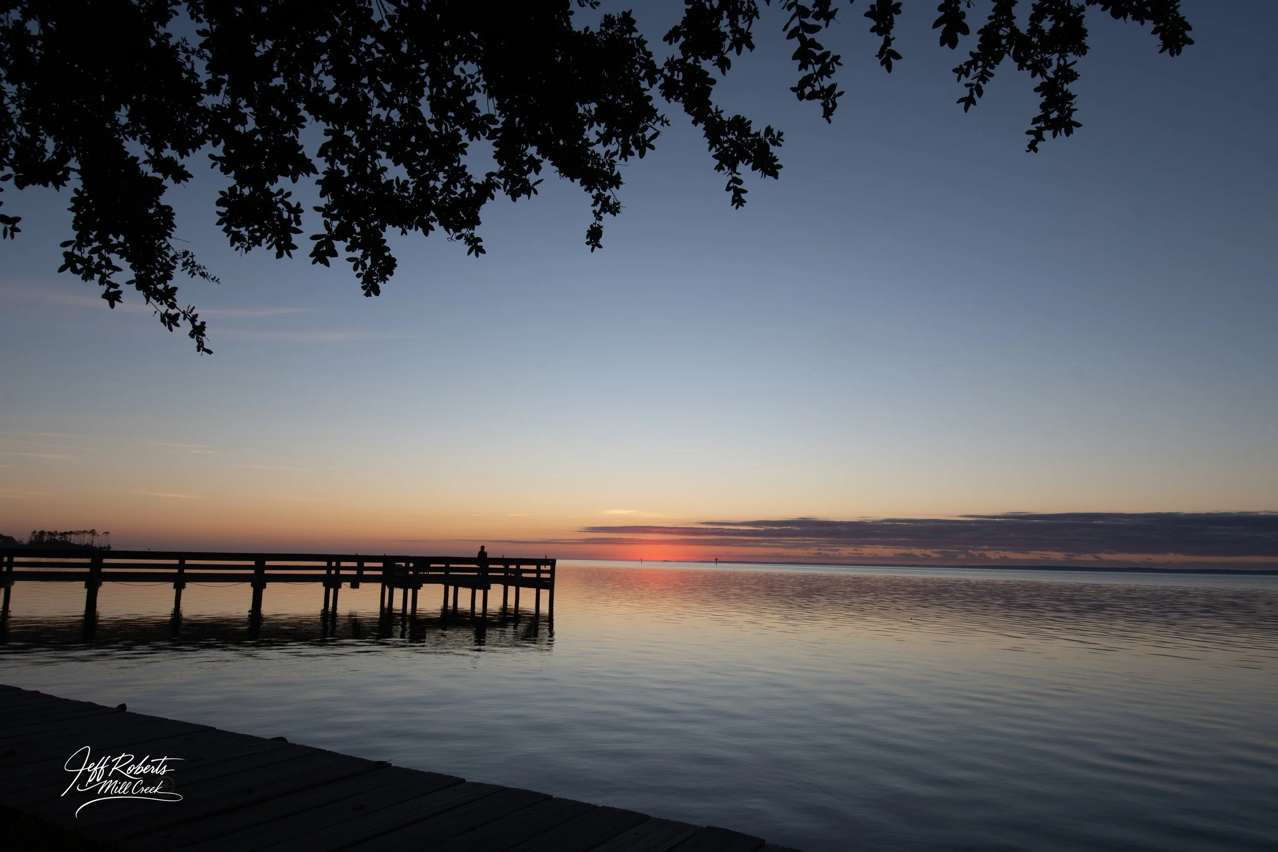 A serene sunset over a calm body of water with a wooden dock extending into it. A person stands on the dock, and tree branches frame the top of the image. The sky is transitioning from orange and pink near the horizon to blue. The scene is peaceful a