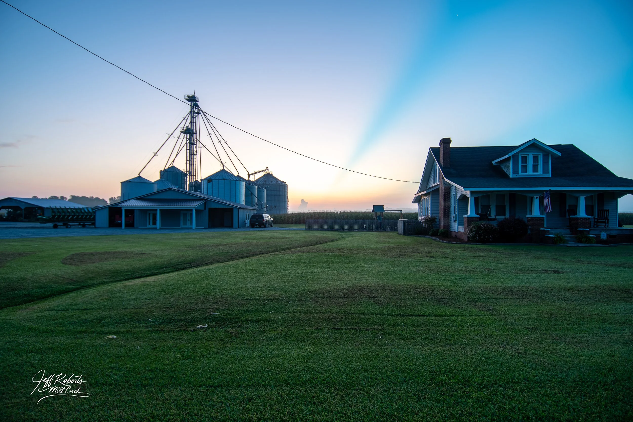 A rural farm scene with a large house on the right and a barn with grain silos and a tall metal grain elevator on the left at sunset or sunrise.