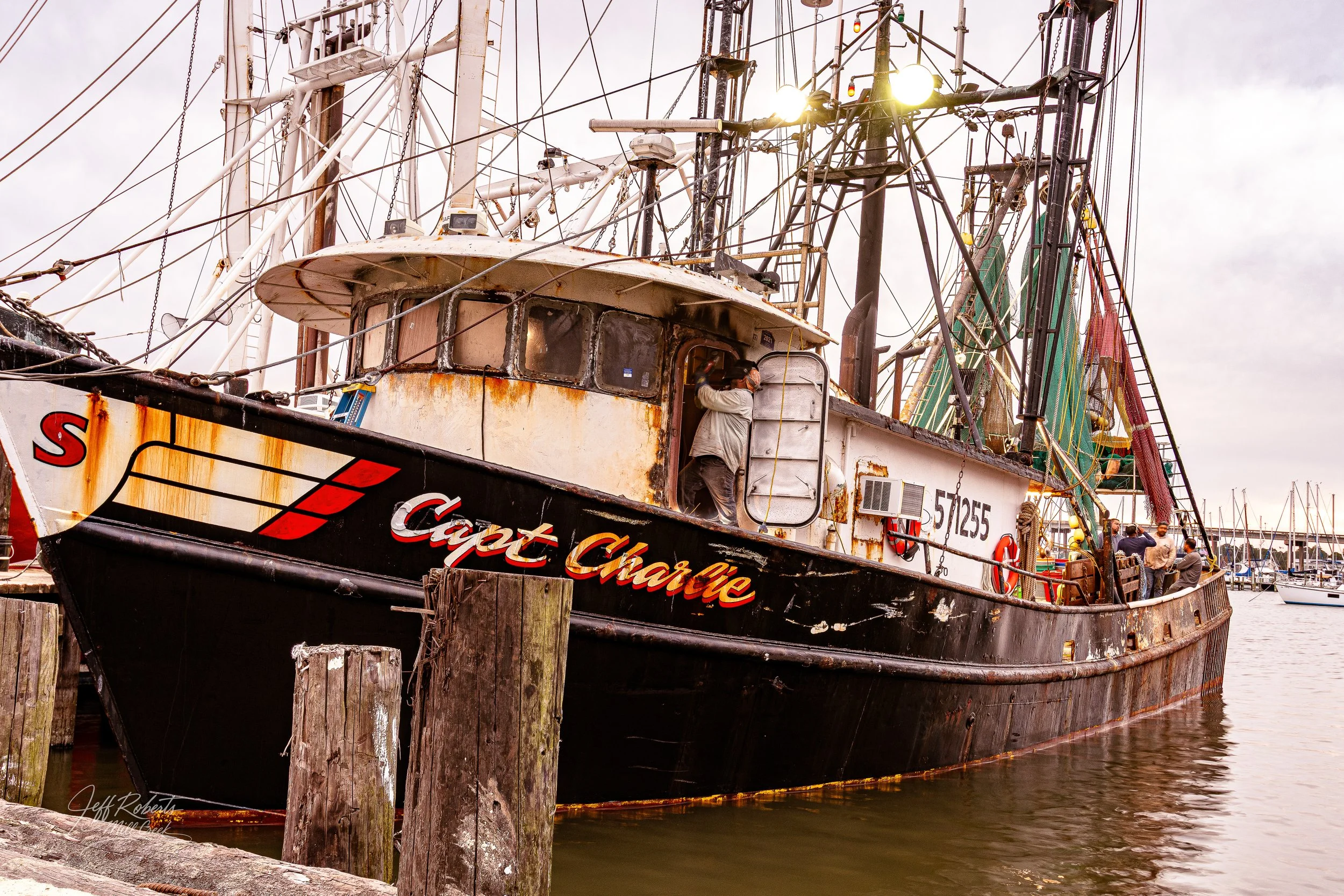 An old, rusted fishing boat named 'Captain Charlie' docked at a marina with other boats in the background. The boat's exterior shows signs of rust and wear, with some people inspecting it.