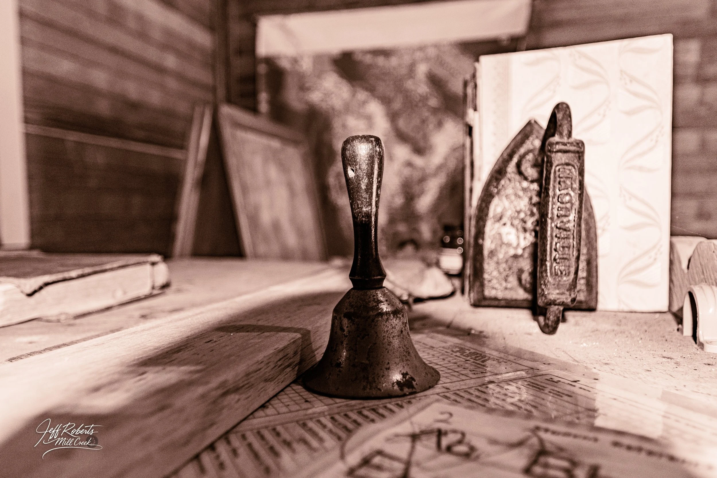 A vintage bell and two metal clamps on a wooden table, with books and a wood-paneled wall in the background.