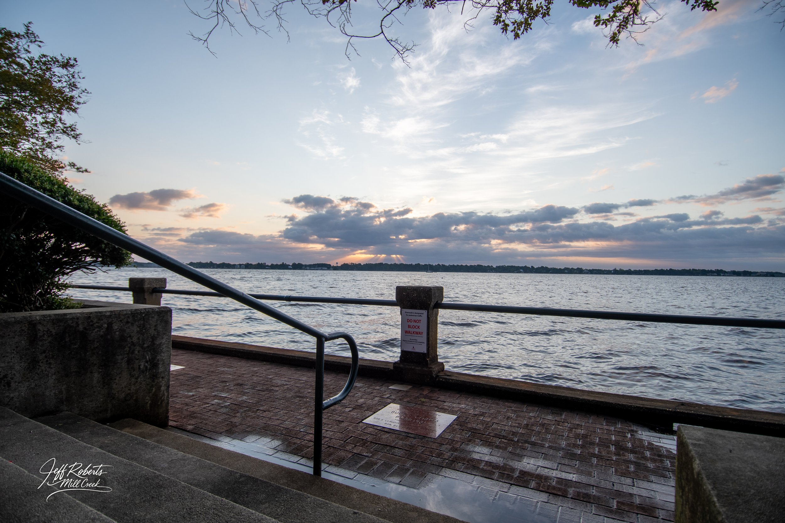 Scenic view of a water body during sunset with clouds in the sky, a walkway with a metal railing, and a sign that reads 'Do Not Block Walkway'.