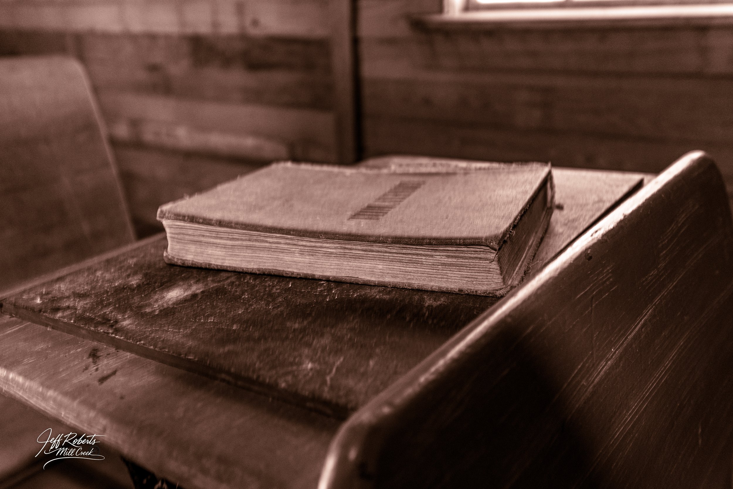 An old, worn book resting on a dark wooden pew inside a rustic, wood-paneled church or chapel.