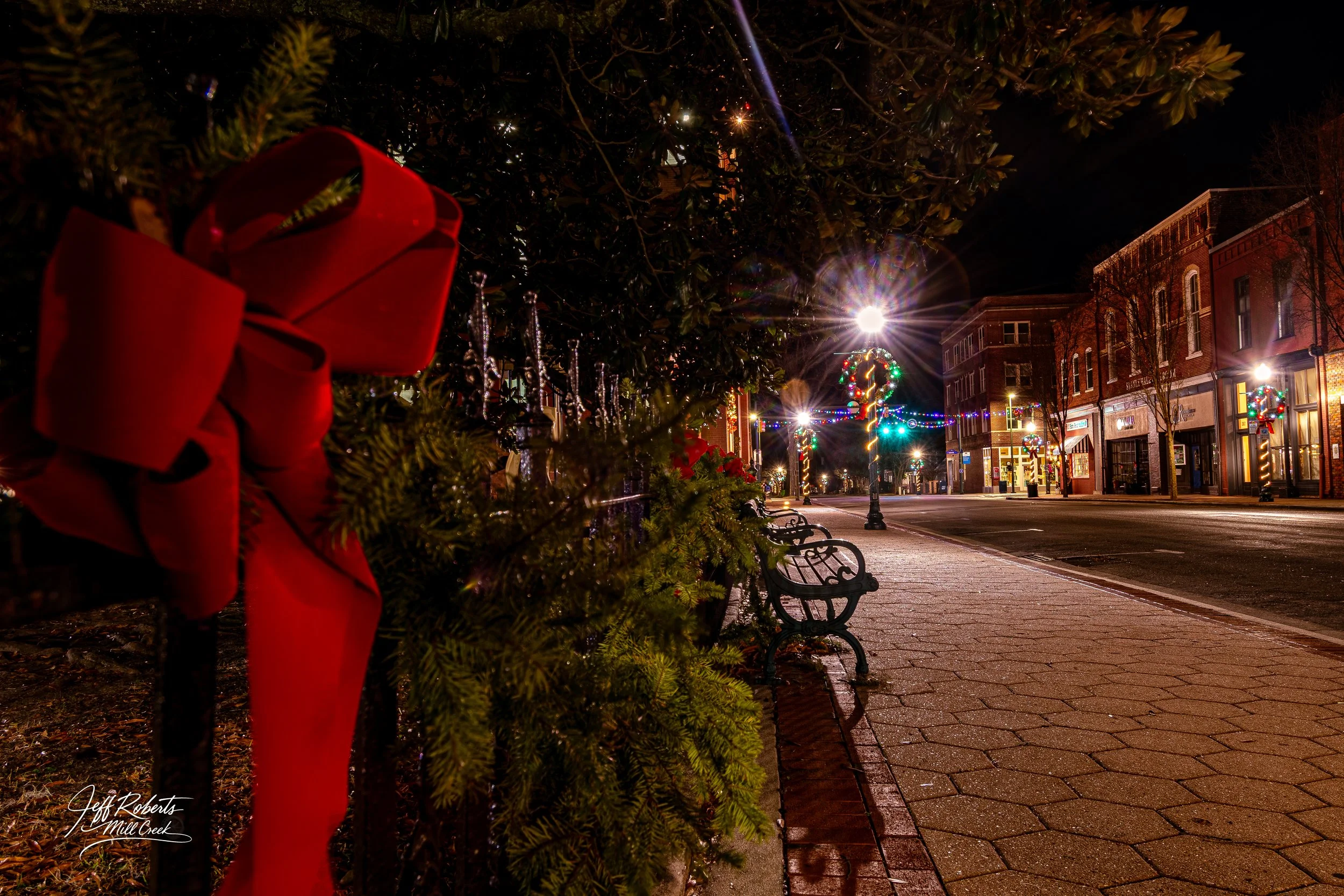 Empty street decorated with Christmas wreaths and lights, with benches along the sidewalk, under streetlights, during nighttime.