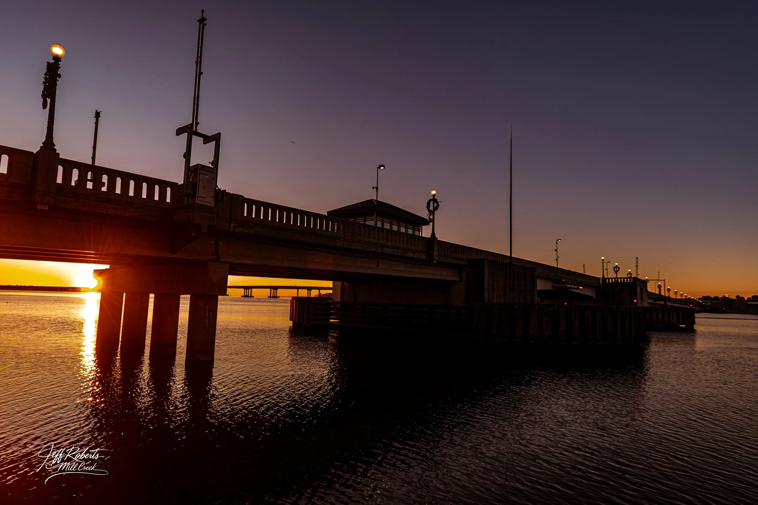 A river bridge at sunset with the sun near the horizon, cast reflections on the water, and street lamps on the bridge.