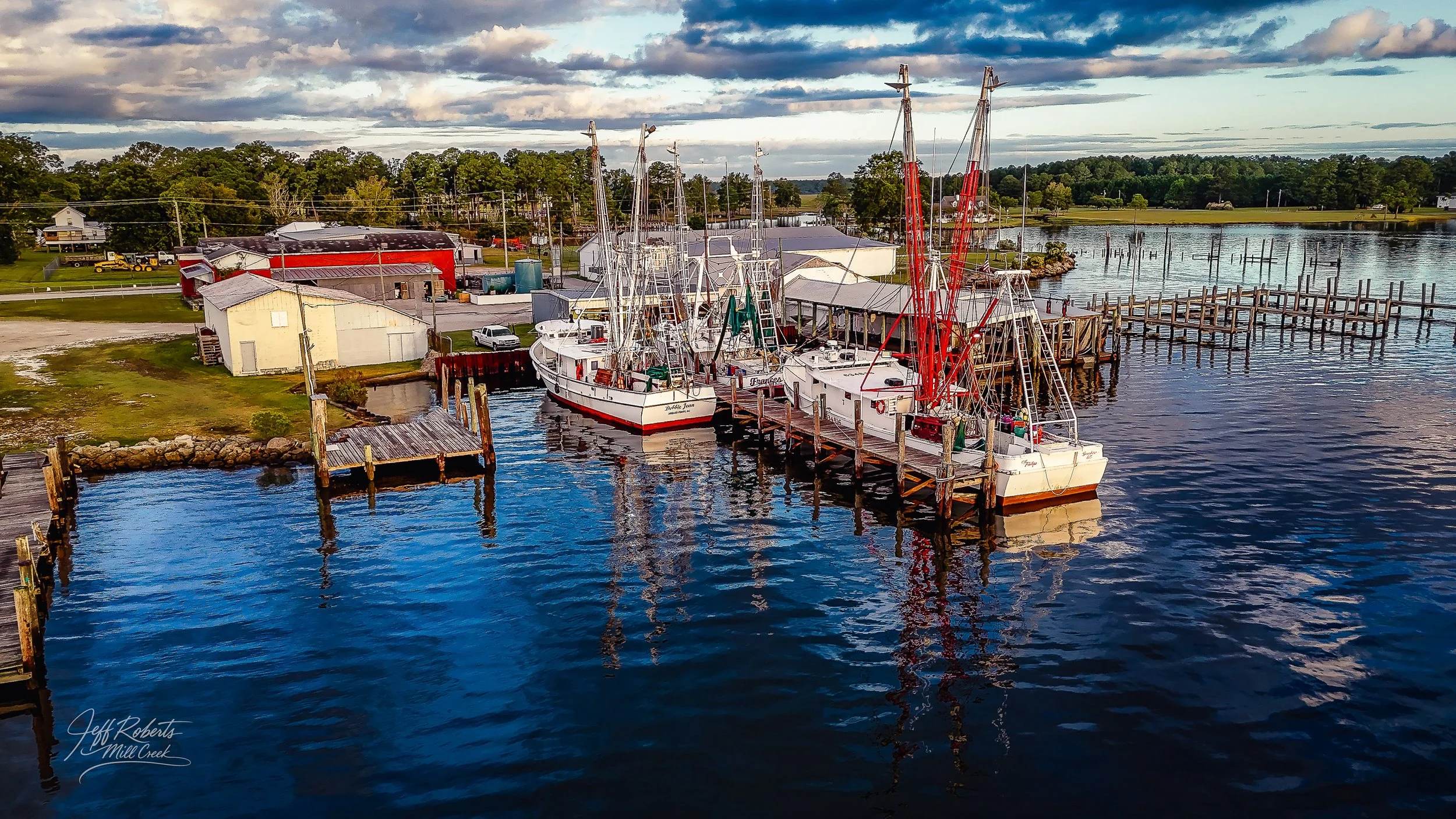 Aerial view of a marina with boats docked at wooden piers and a small boatyard, with buildings, boats, and trees in the background under a cloudy sky.
