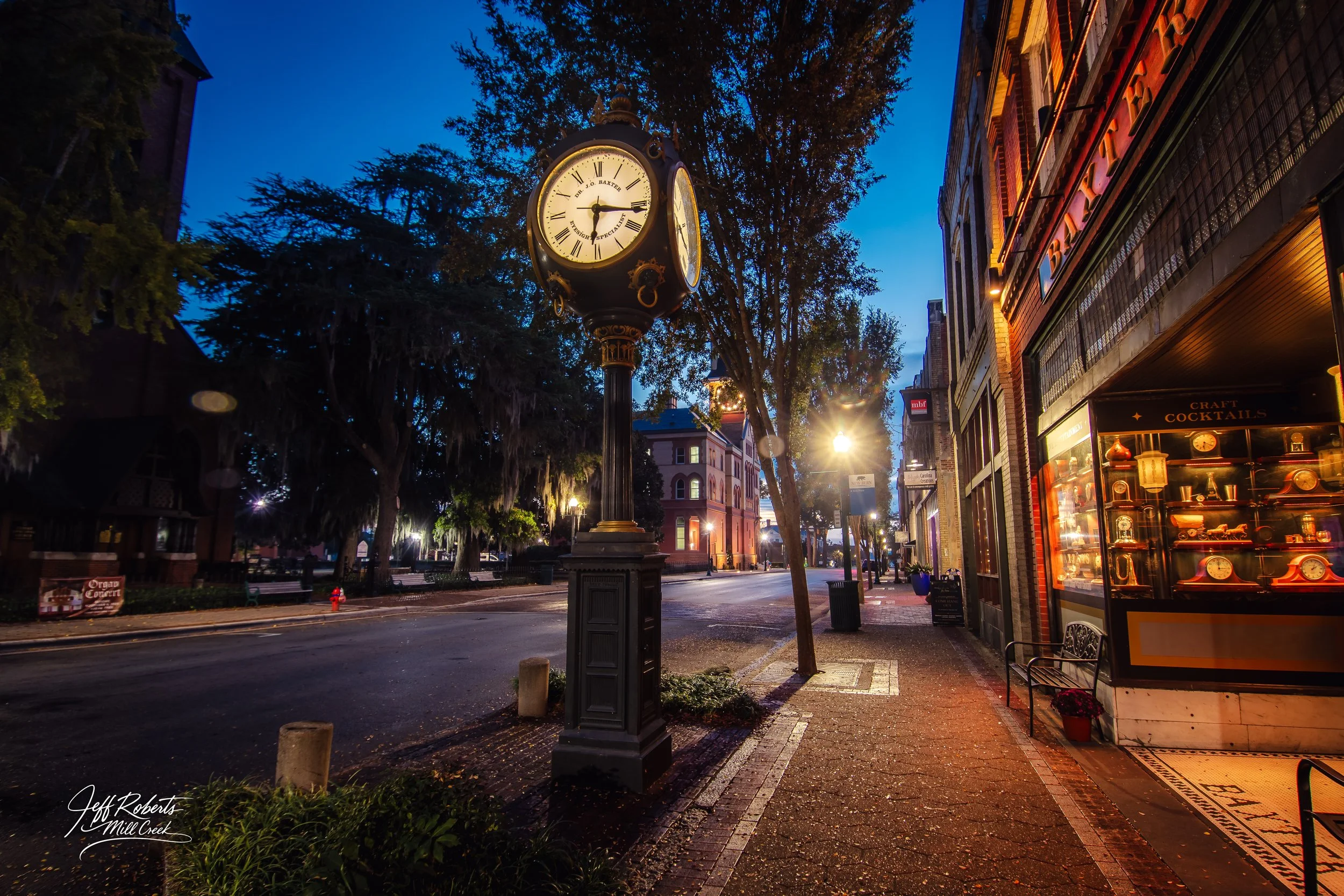 Downtown street scene at dusk with an illuminated clock on a pole, trees lining the sidewalk, storefronts including a craft cocktail shop, and streetlights casting a warm glow.