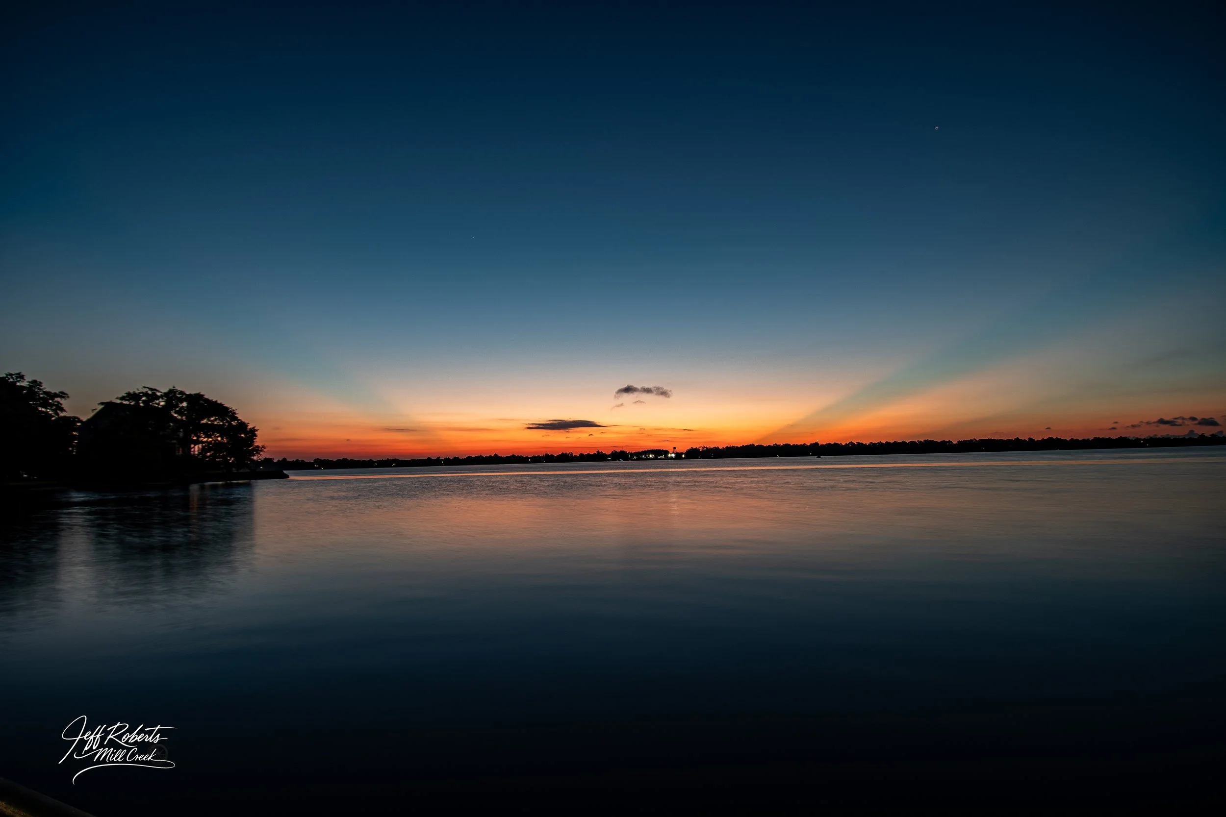 A peaceful sunset over a large body of water with a silhouetted shoreline and trees on the left side, colorful sky with gradients from orange to deep blue.