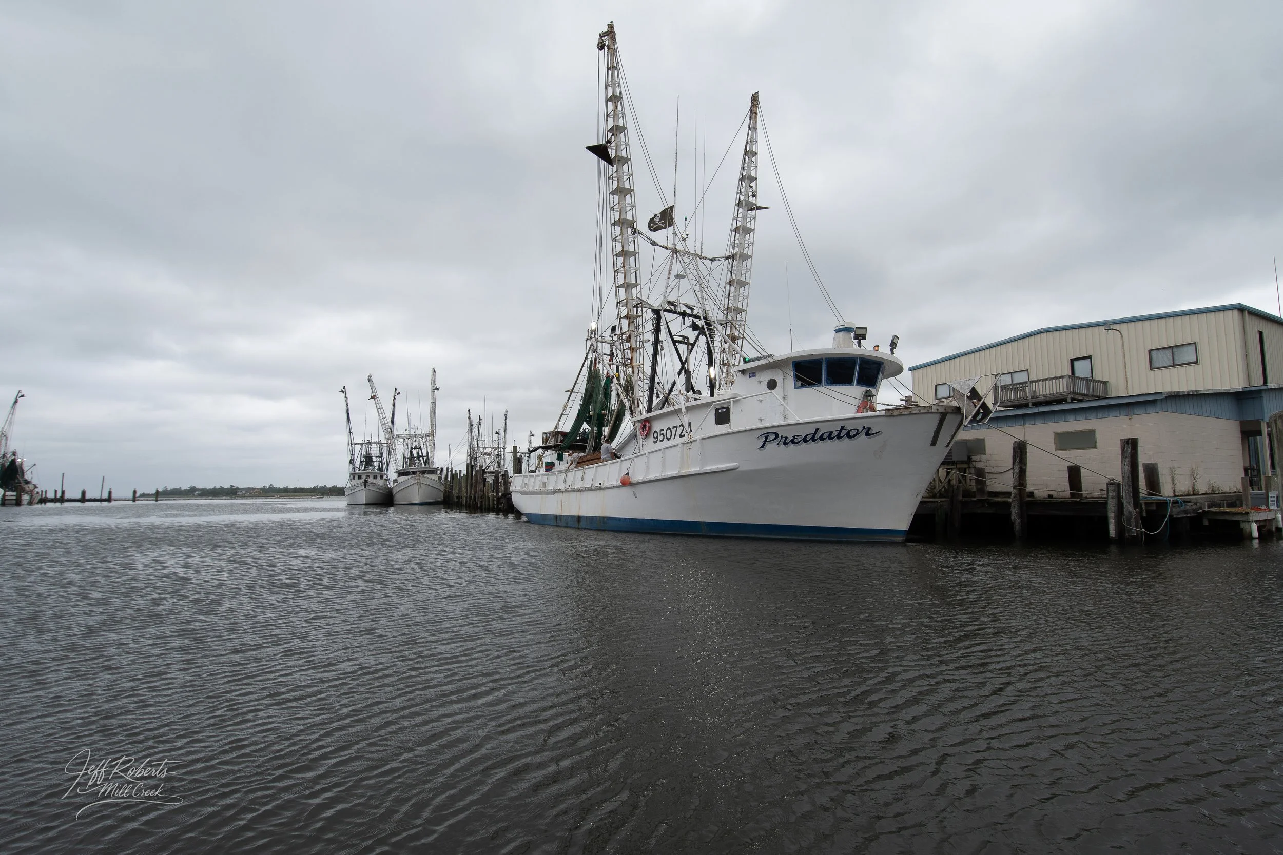 Several fishing boats docked at a marina on a cloudy day, with a building on the right side.