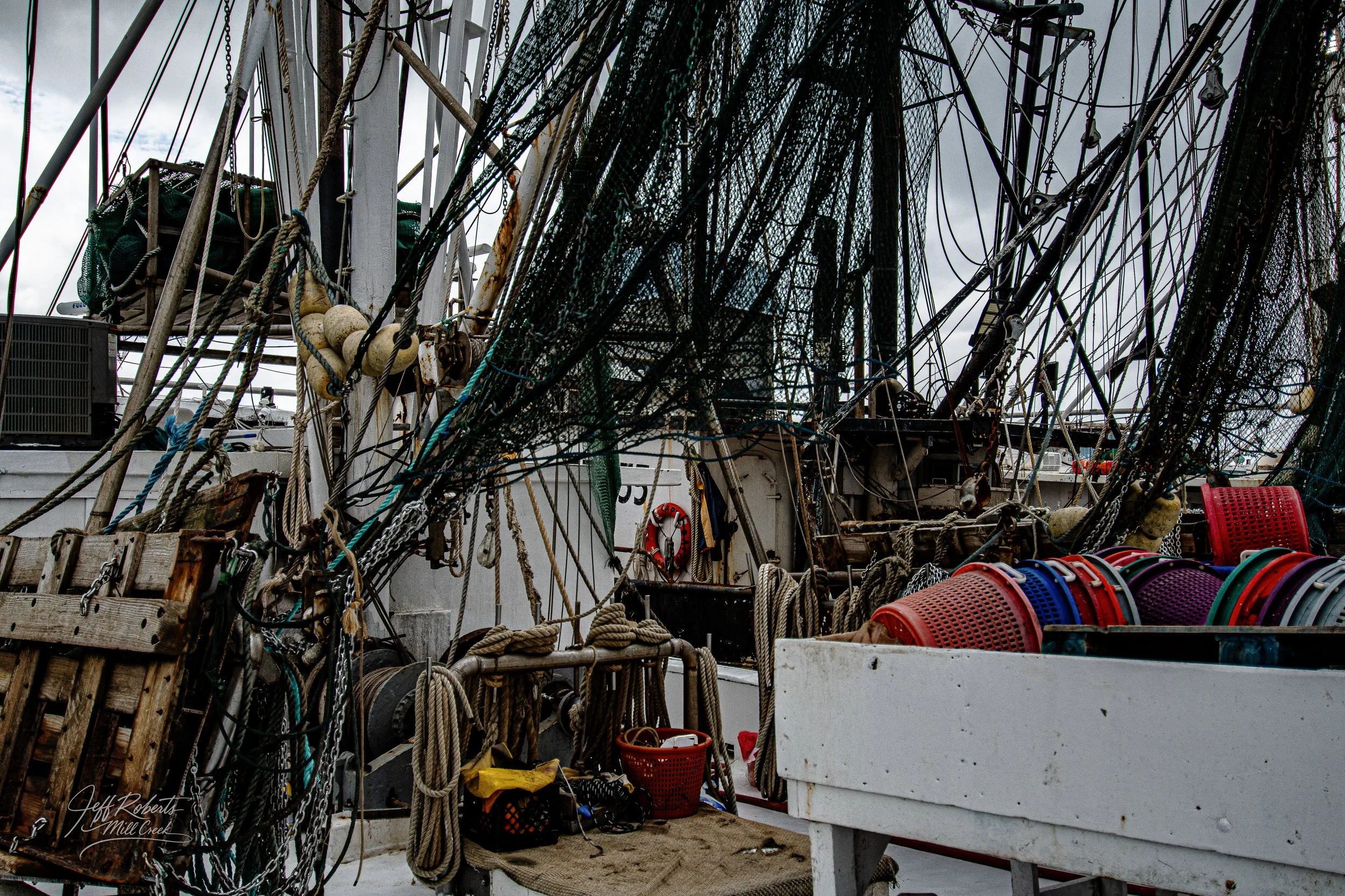 Close-up view of a fishing boat deck featuring tangled ropes, nets, and colorful plastic buckets, with a cloudy sky overhead.