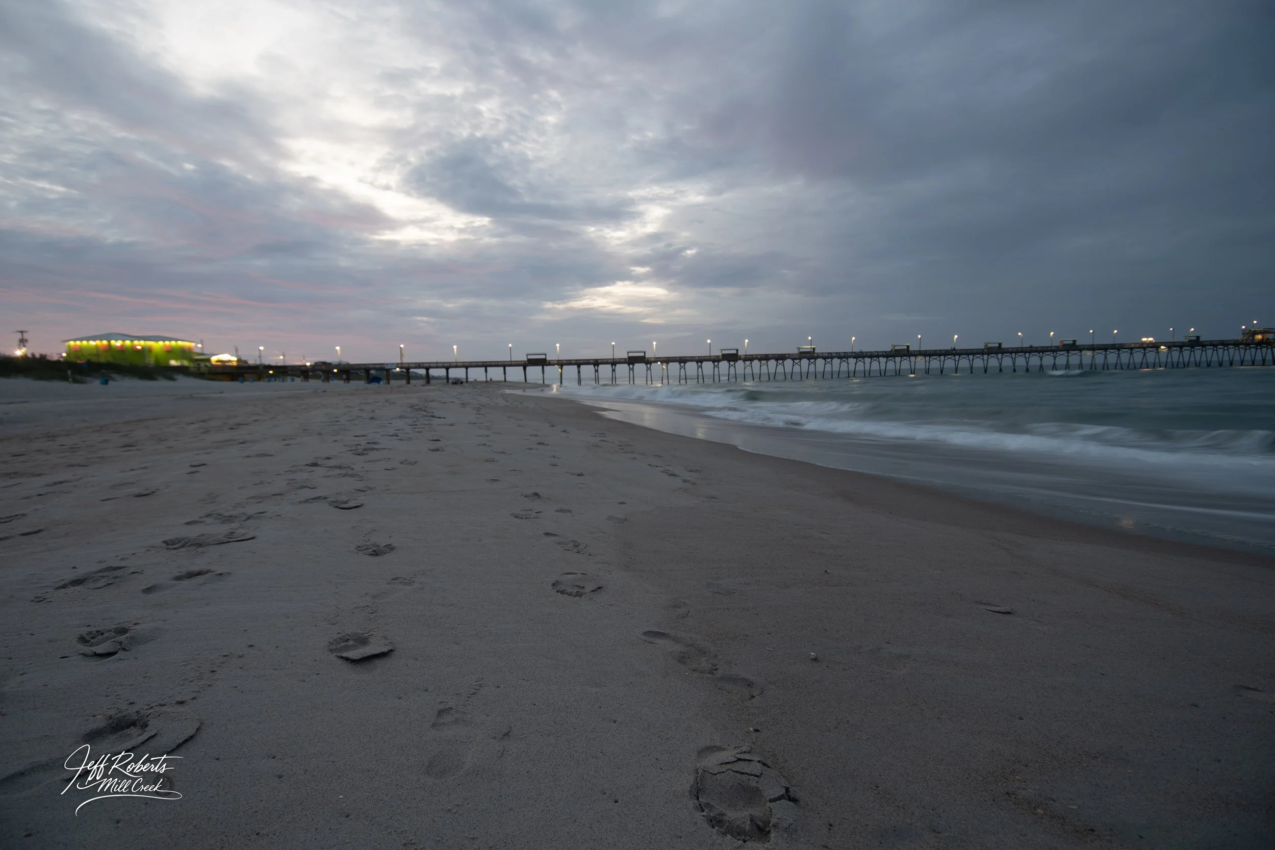 Footprints in the sand on a beach with a pier extending into the ocean under a cloudy sky during twilight.