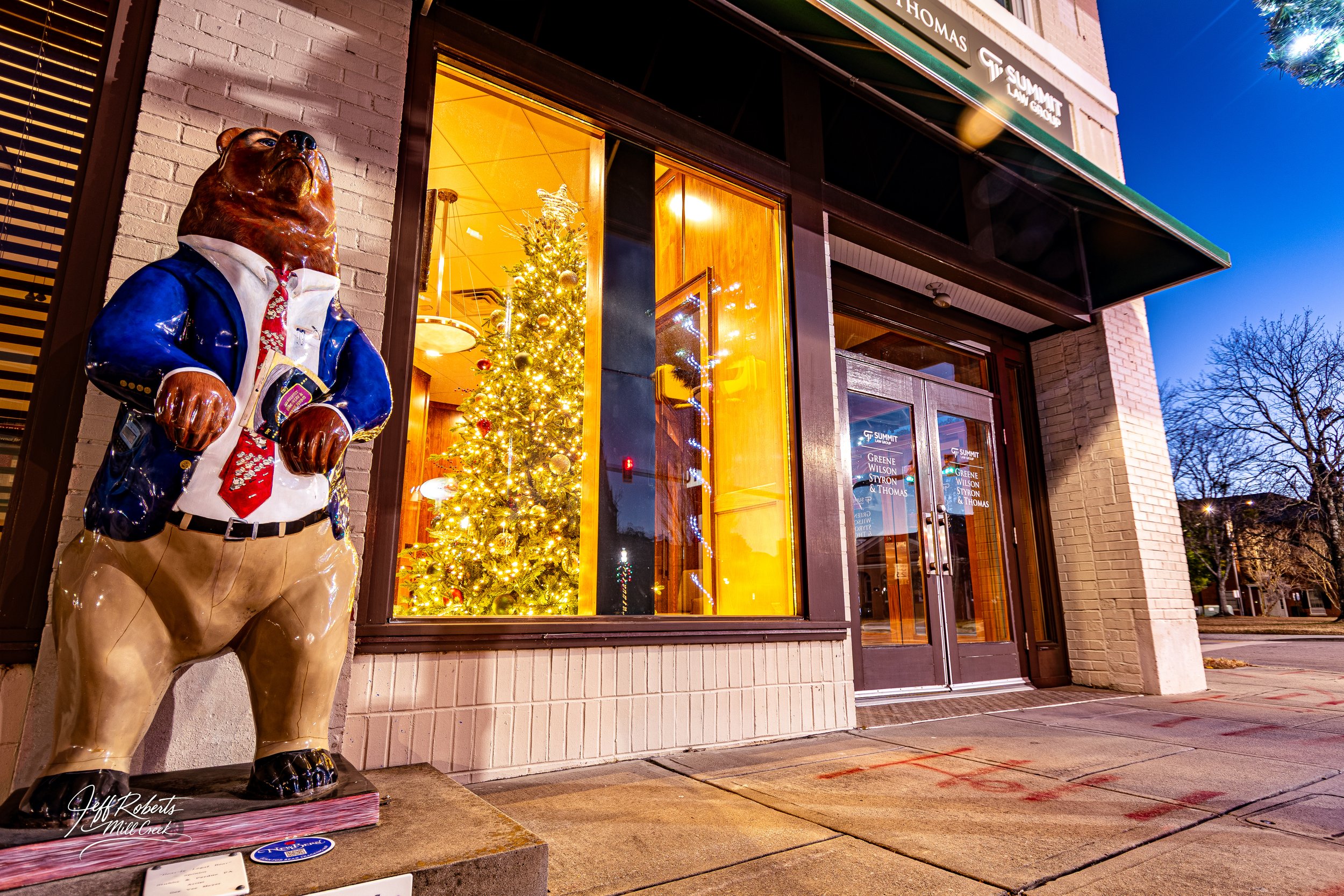 A bear statue dressed in a suit with a festive tie stands outside a building during the evening. The building has large windows with a decorated Christmas tree lit inside and the street is visible in the background.