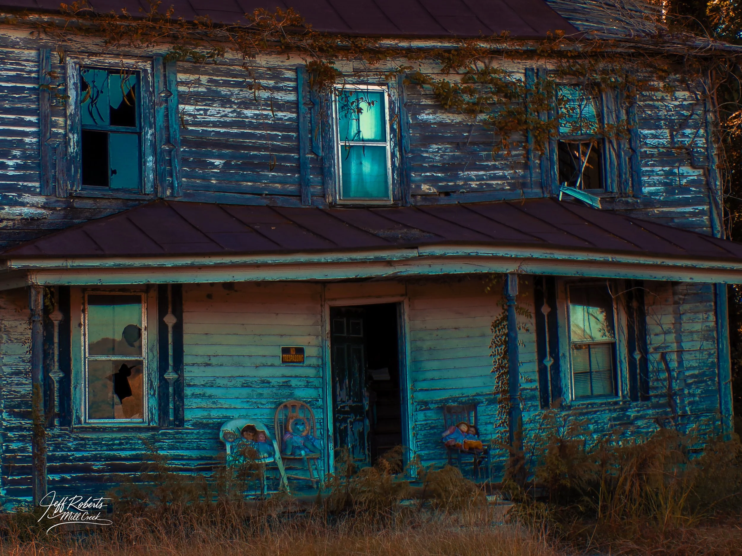 An abandoned, weathered two-story house with peeling blue and white paint, broken windows, and overgrown bushes. The front porch has old dolls and a chair, with vines growing along the exterior.