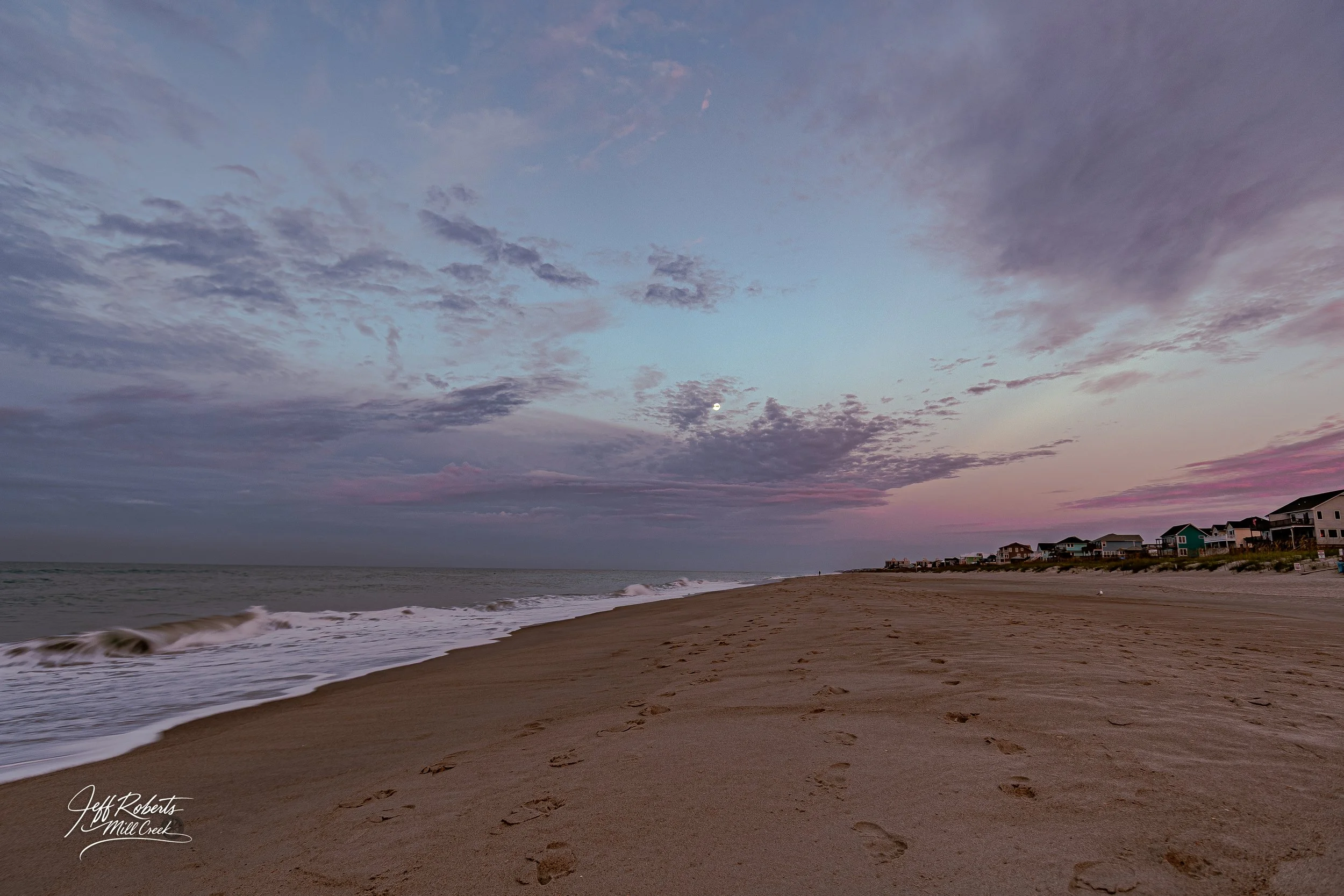 Beach at sunset with footprints in the sand, ocean waves, houses along the shoreline, cloudy sky with the moon visible.