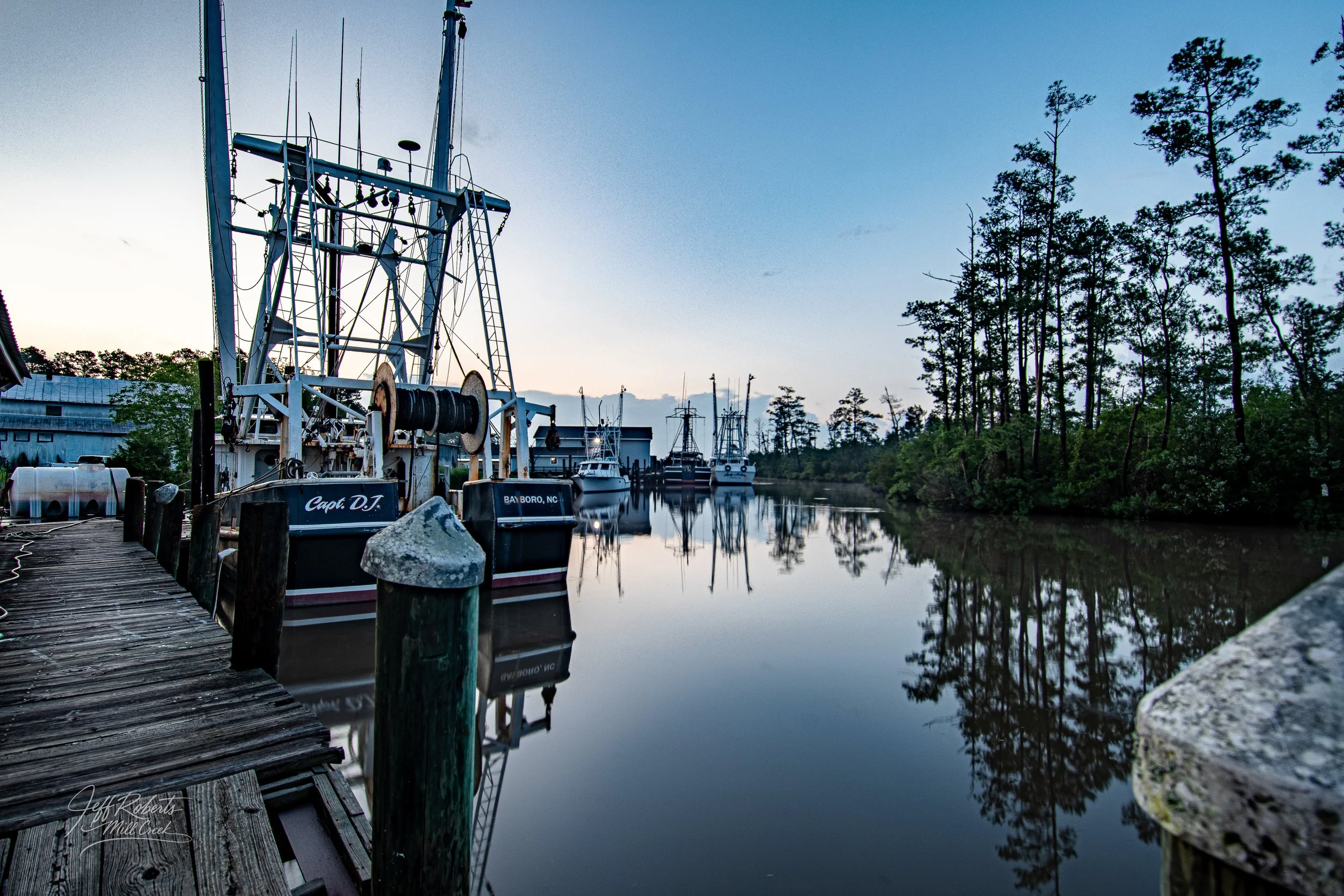 View of boats docked along a wooden pier on a calm waterway, with trees and a blue sky in the background.