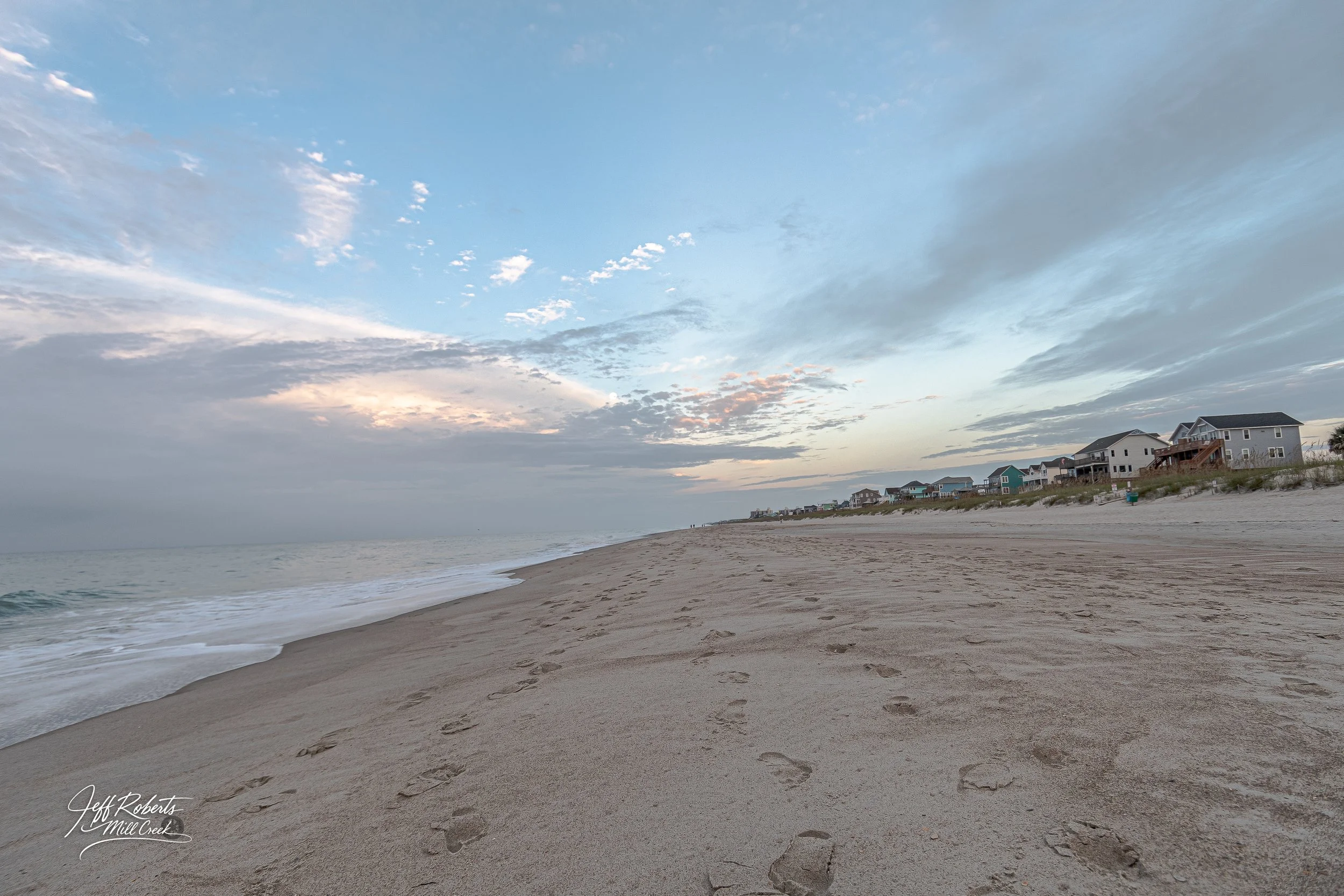 A sandy beach with footprints, ocean waves, and a line of beach houses under a partly cloudy sky at dusk.