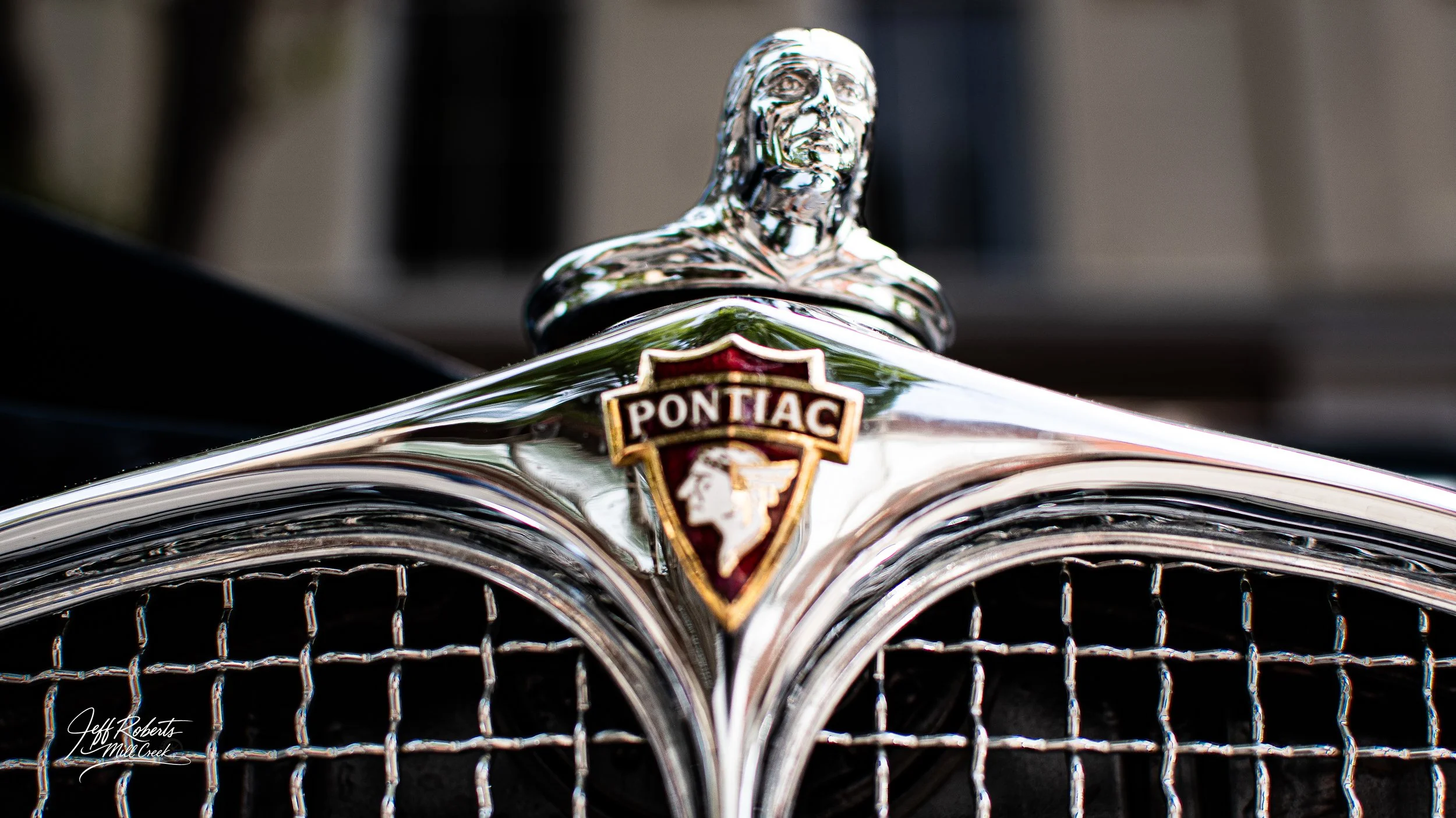 Close-up of a vintage Pontiac car hood ornament and grille, featuring a chrome statue of a Native American figure and the Pontiac emblem with a shield shape and profile of a Native American chief.