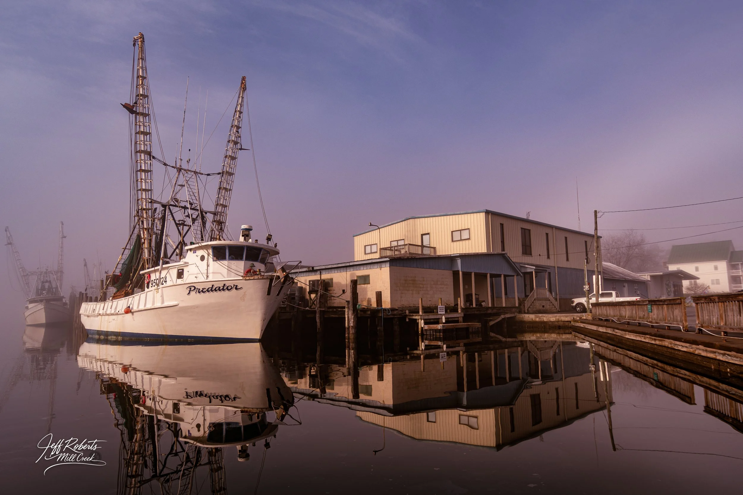 A boat named Predator docked by a building on a calm water reflective surface, with foggy background and other boats visible.