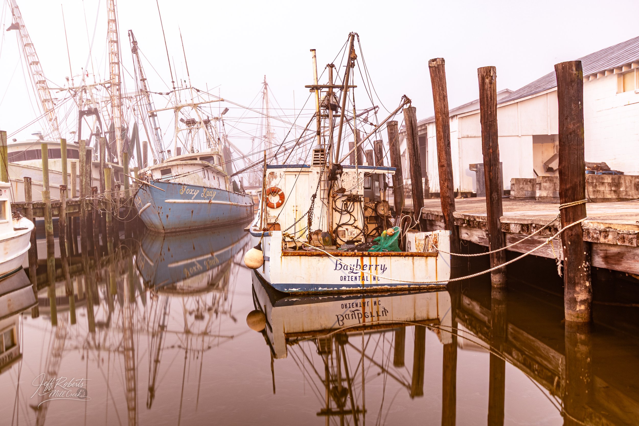 Old fishing boats docked at a pier with calm water reflecting the boats and the wooden pilings, a white building in the background.