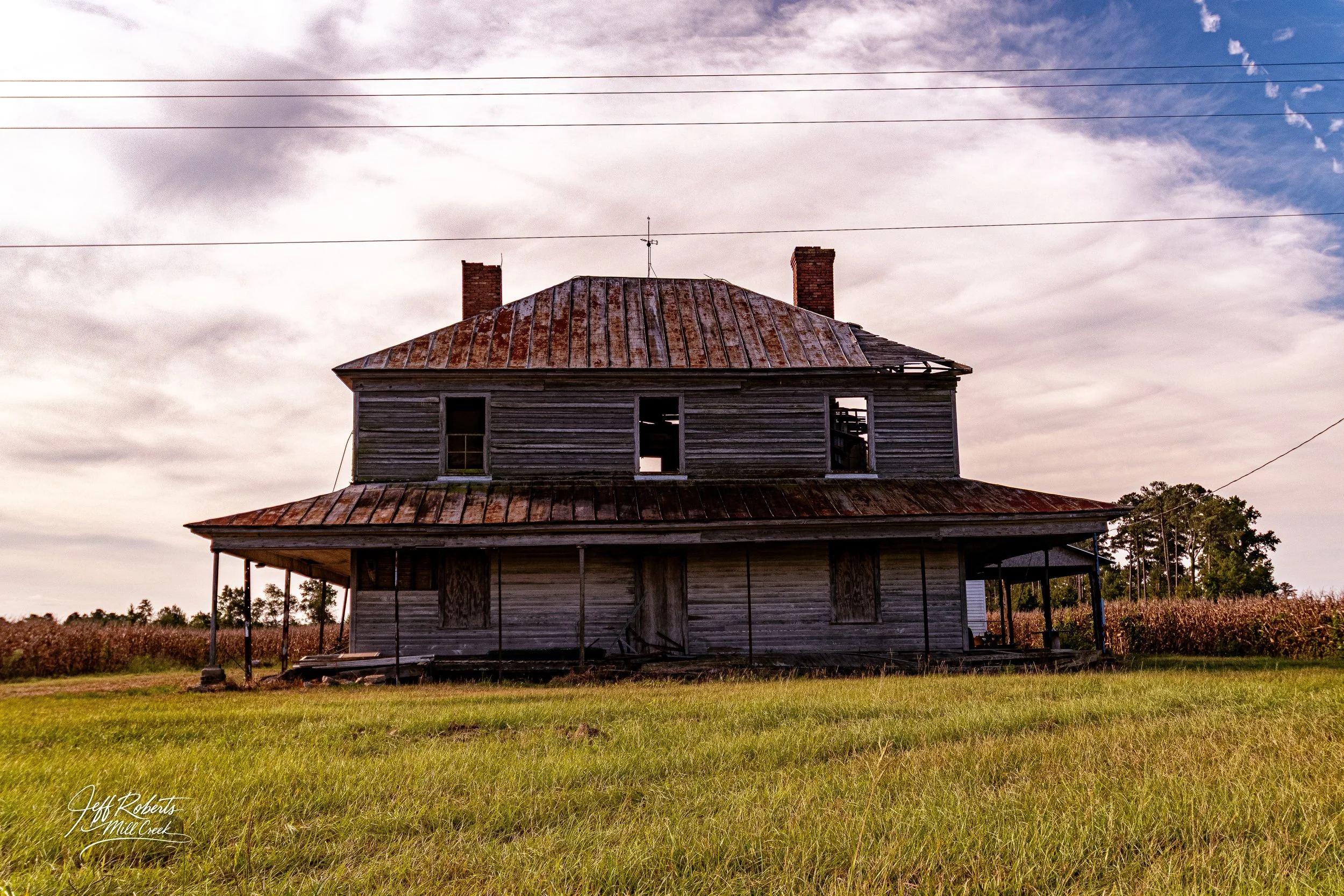 A weathered, abandoned two-story wooden house with a rusted metal roof, set in a grassy field with a field of crops in the background under a partly cloudy sky.