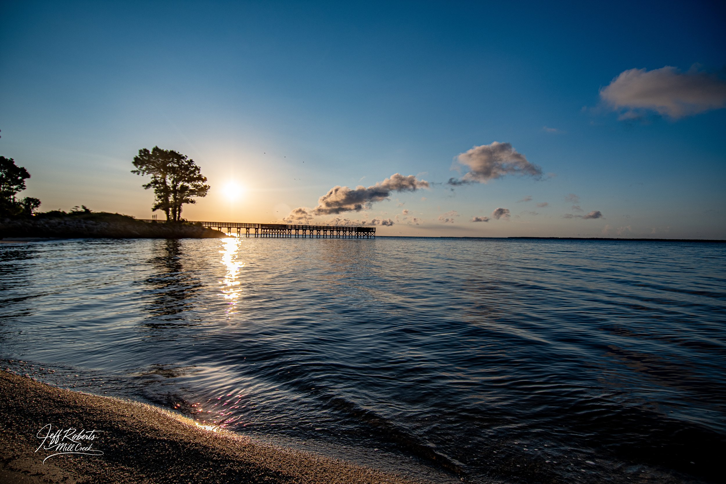 Sunset over a calm body of water with a wooden pier and trees on the shore.