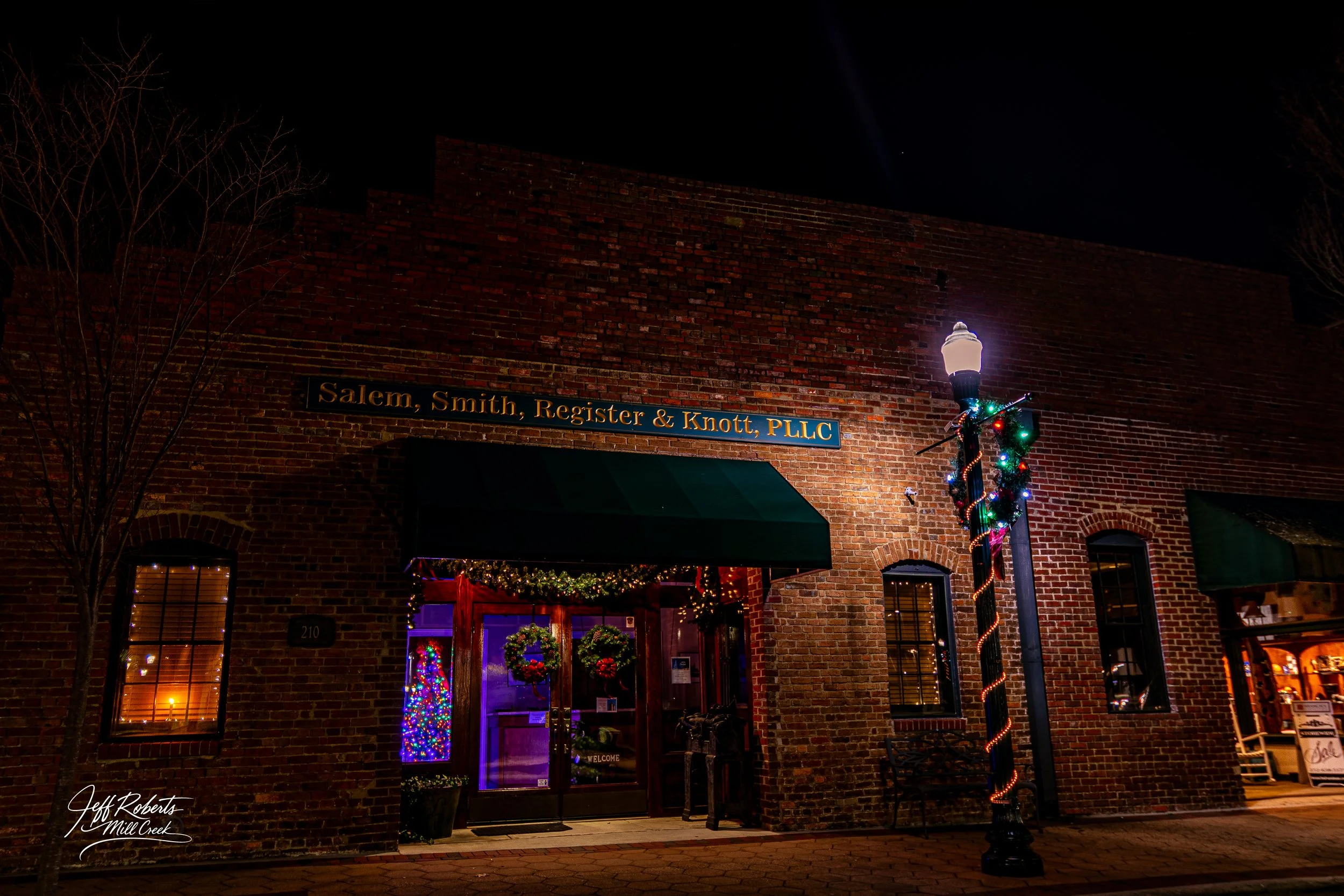 Nighttime view of a brick building with a sign that reads 'Salem, Smith, Register & Knott, PLLC'. The building has windows illuminated from inside, festive Christmas decorations including wreaths and colorful lights, and a lamp post wrapped with Chri