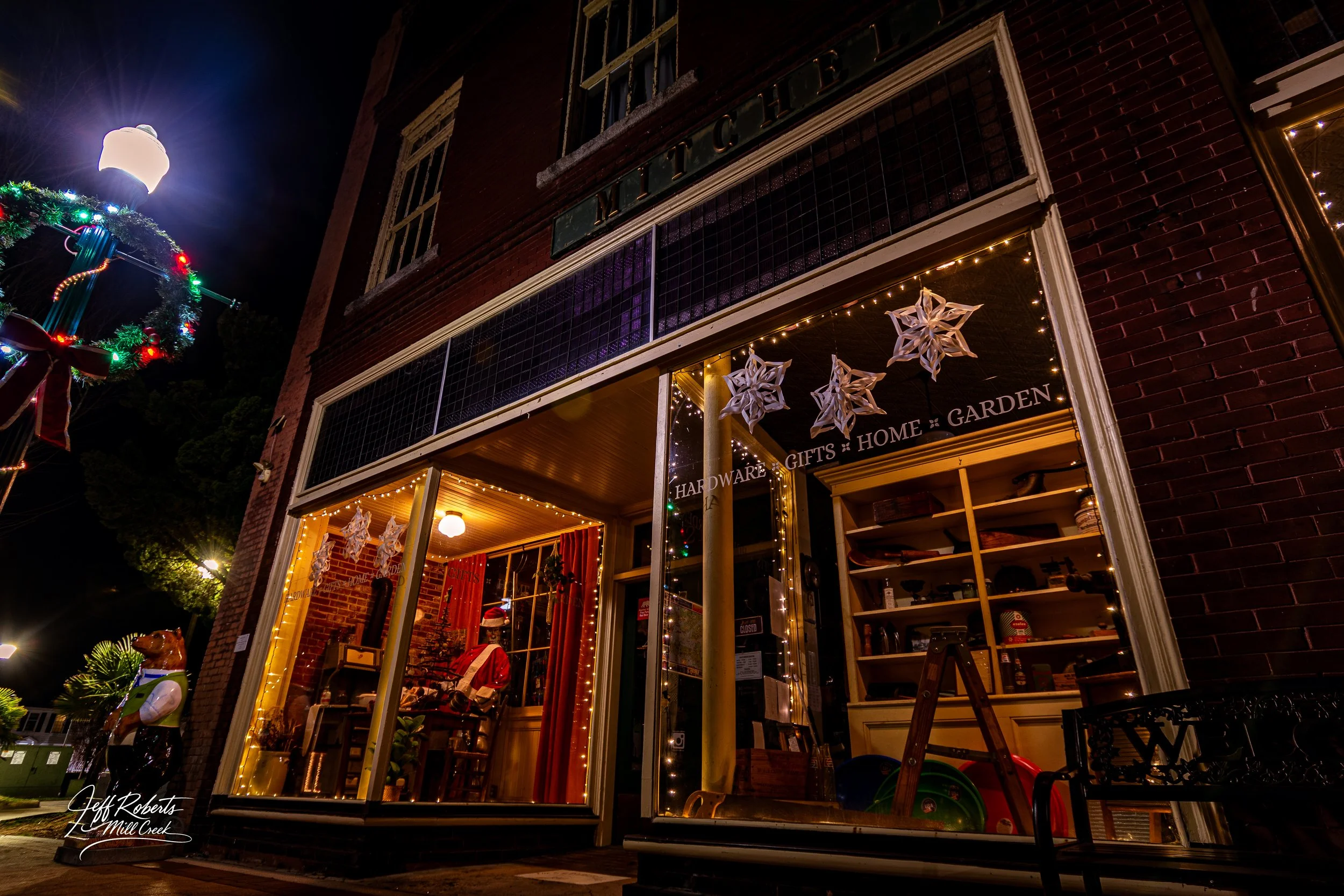 Night view of a decorated storefront with Christmas ornaments, snowflake decorations, and holiday lighting, featuring an inside display with a Santa figure and a bear statue outside.