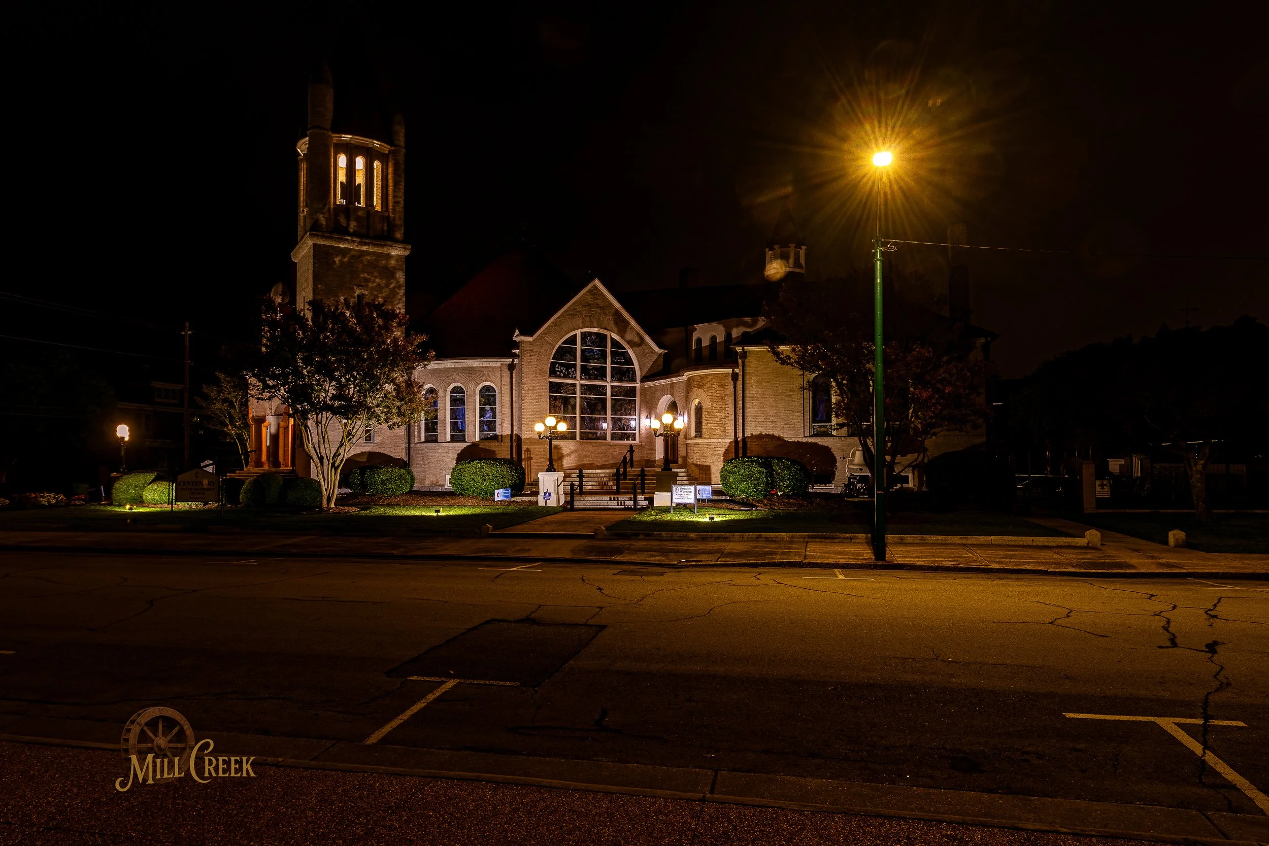 Night view of a well-lit church with a tall tower, surrounded by trees and bushes, streetlights, and a parking lot in the foreground.