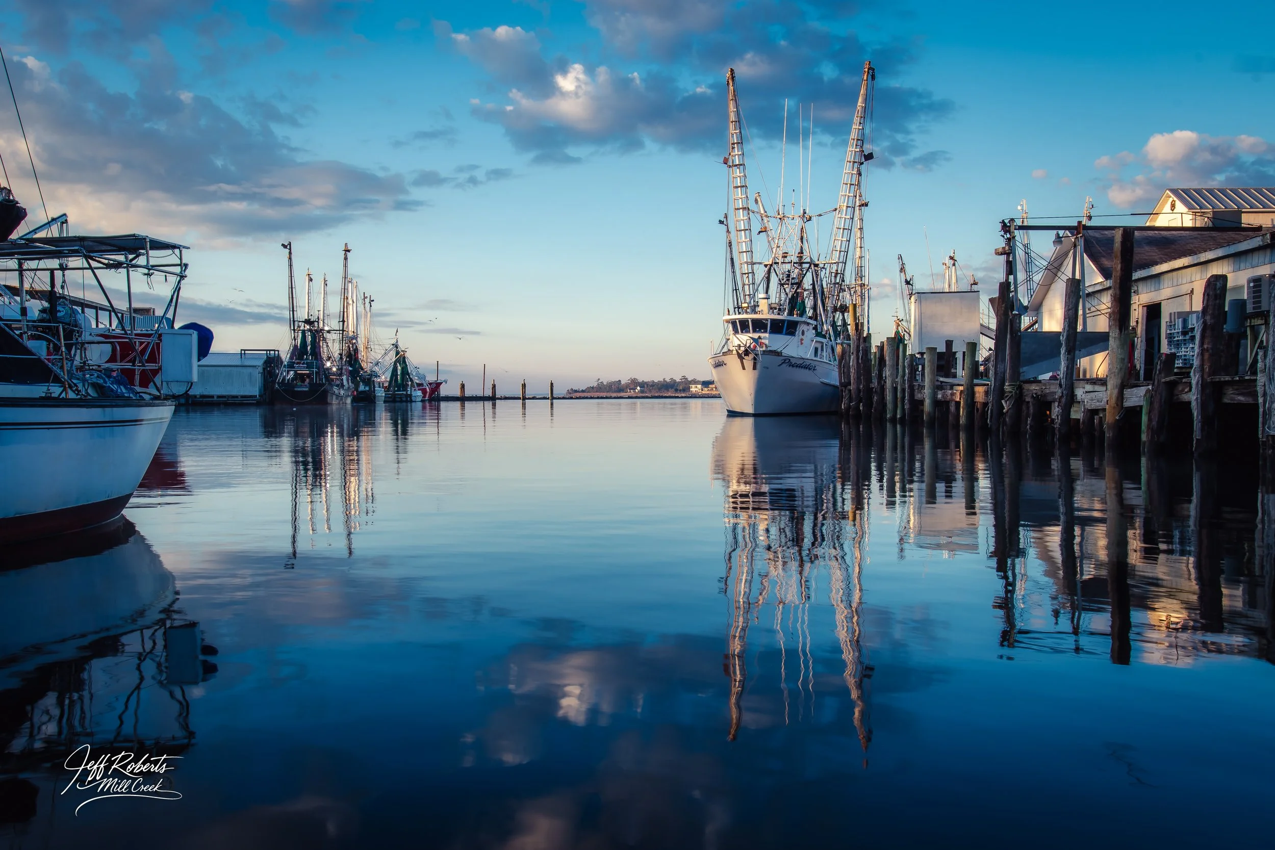 Marina with sailboats and docked boats on calm water during sunset, with a partly cloudy sky.