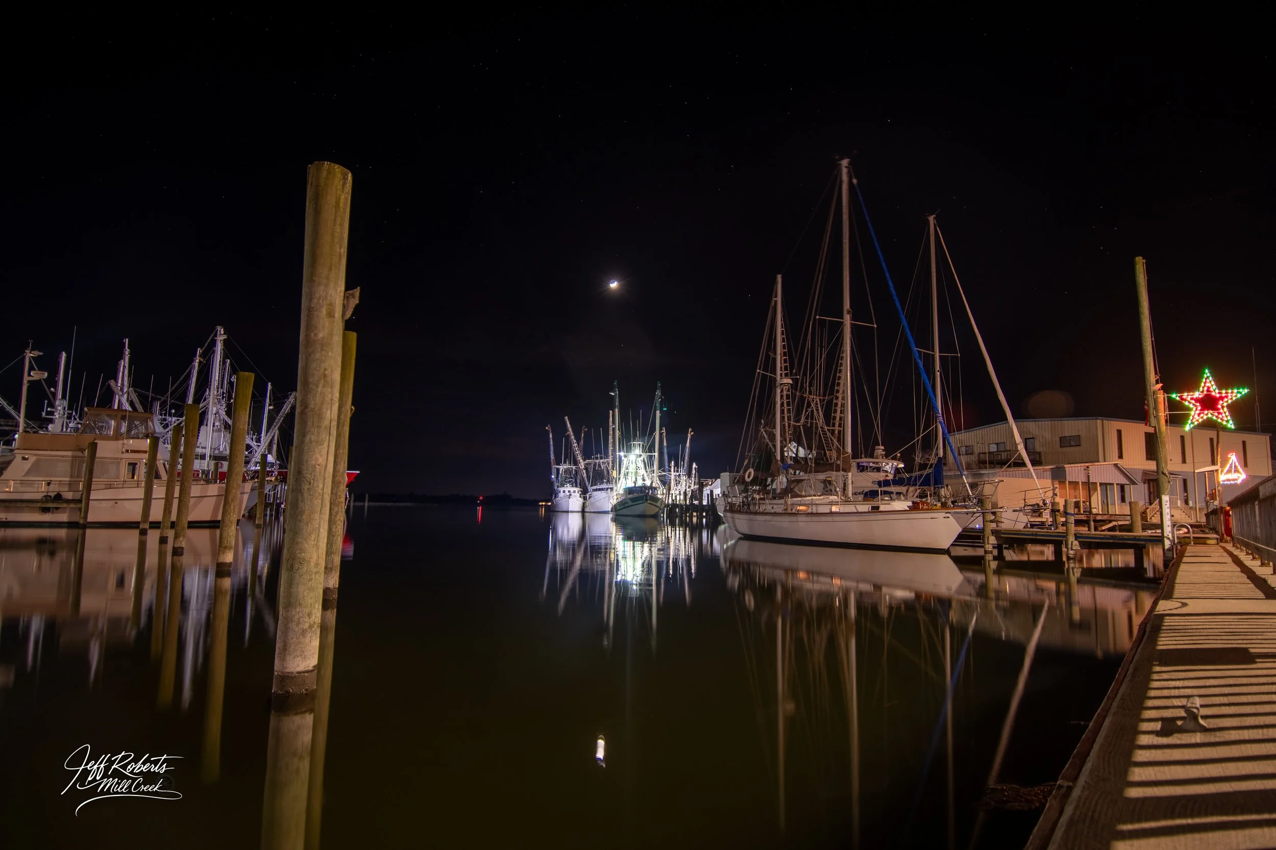 Night view of a marina with boats docked and their reflections in calm water, under a starry sky with a crescent moon, and festive Christmas lights on a nearby building.