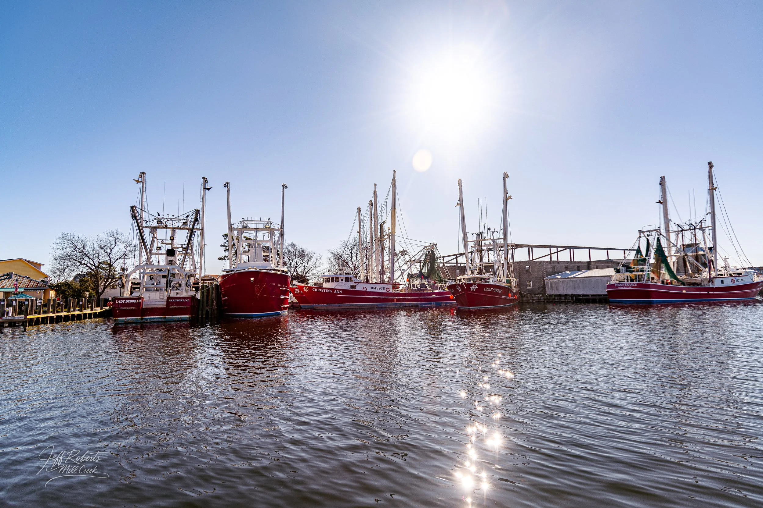 Six boats docked along a pier in a marina with the sun shining brightly overhead and reflecting on the water.