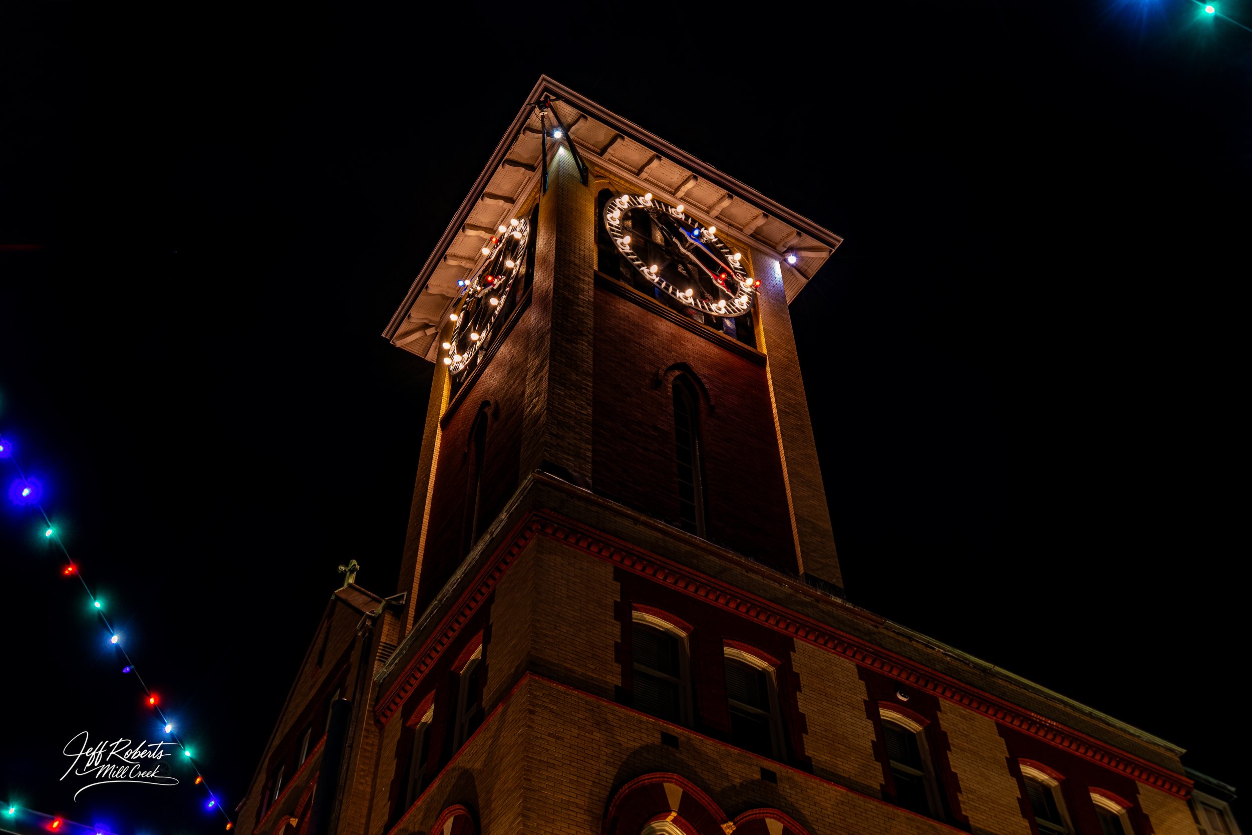 Nighttime photo of a tall brick clock tower with lit clock faces, illuminated edges, and colorful string lights hanging nearby.