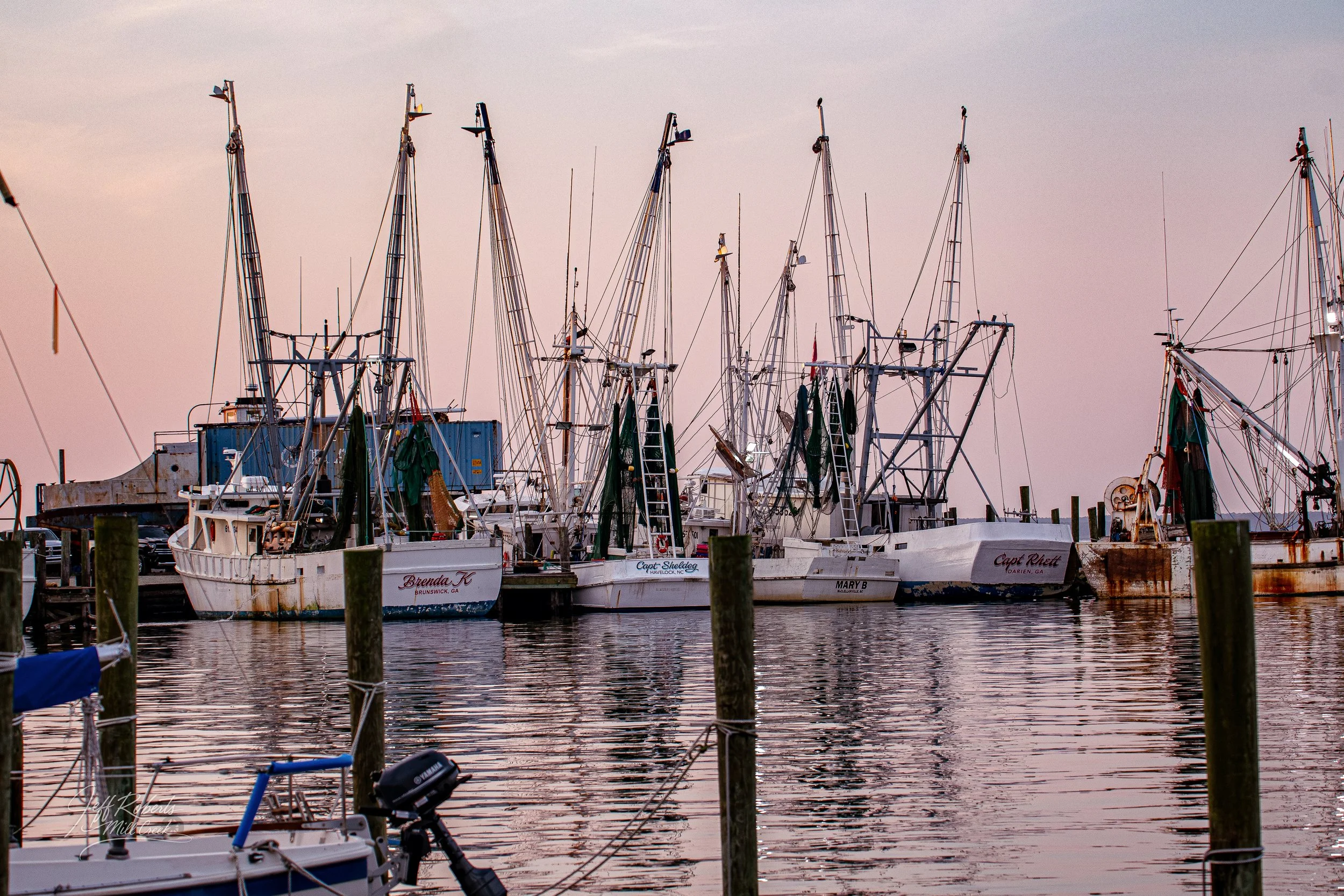 A marina filled with docked sailboats and fishing boats at sunset, with water reflecting the pastel sky.