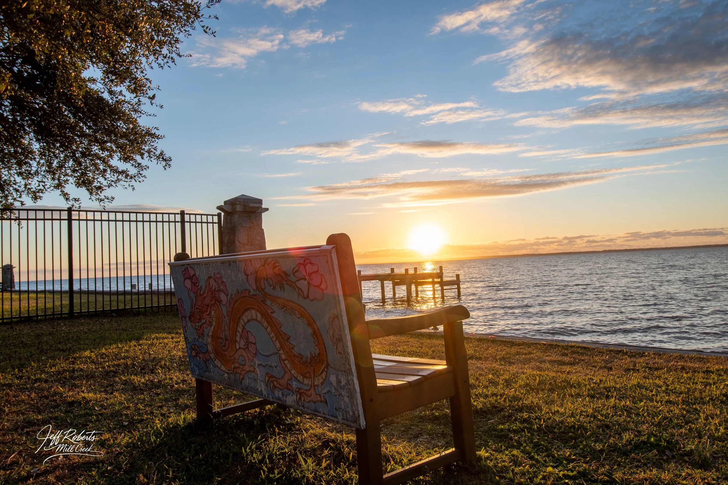 A wooden bench with a colorful dragon design on its backrest faces a lakeside at sunset. The scene includes a fence, a stone pillar, a small pier extending into the water, and a partly cloudy sky with the sun near the horizon.