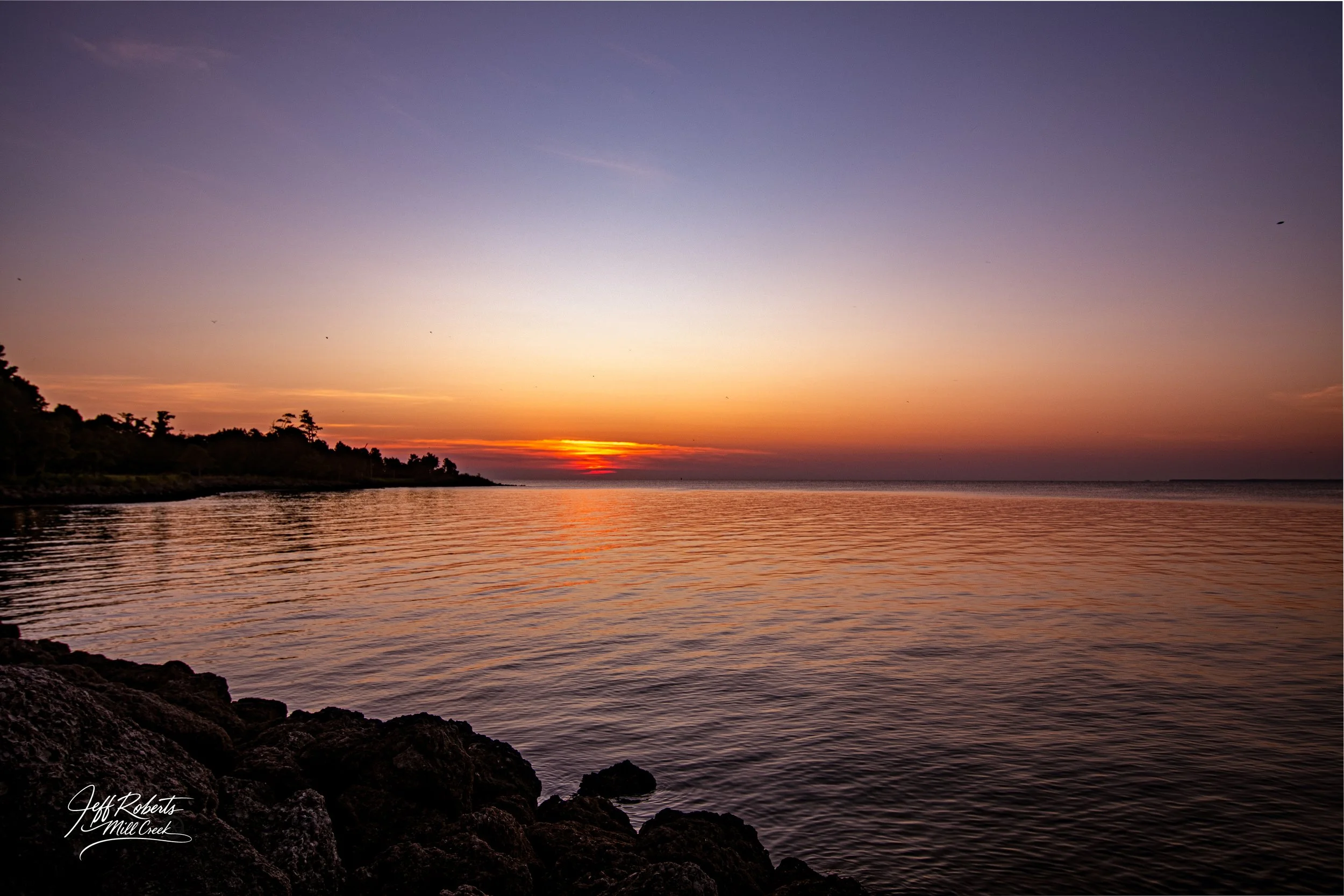 Sunset over a calm body of water with rocky shoreline in the foreground and silhouettes of trees on the horizon.