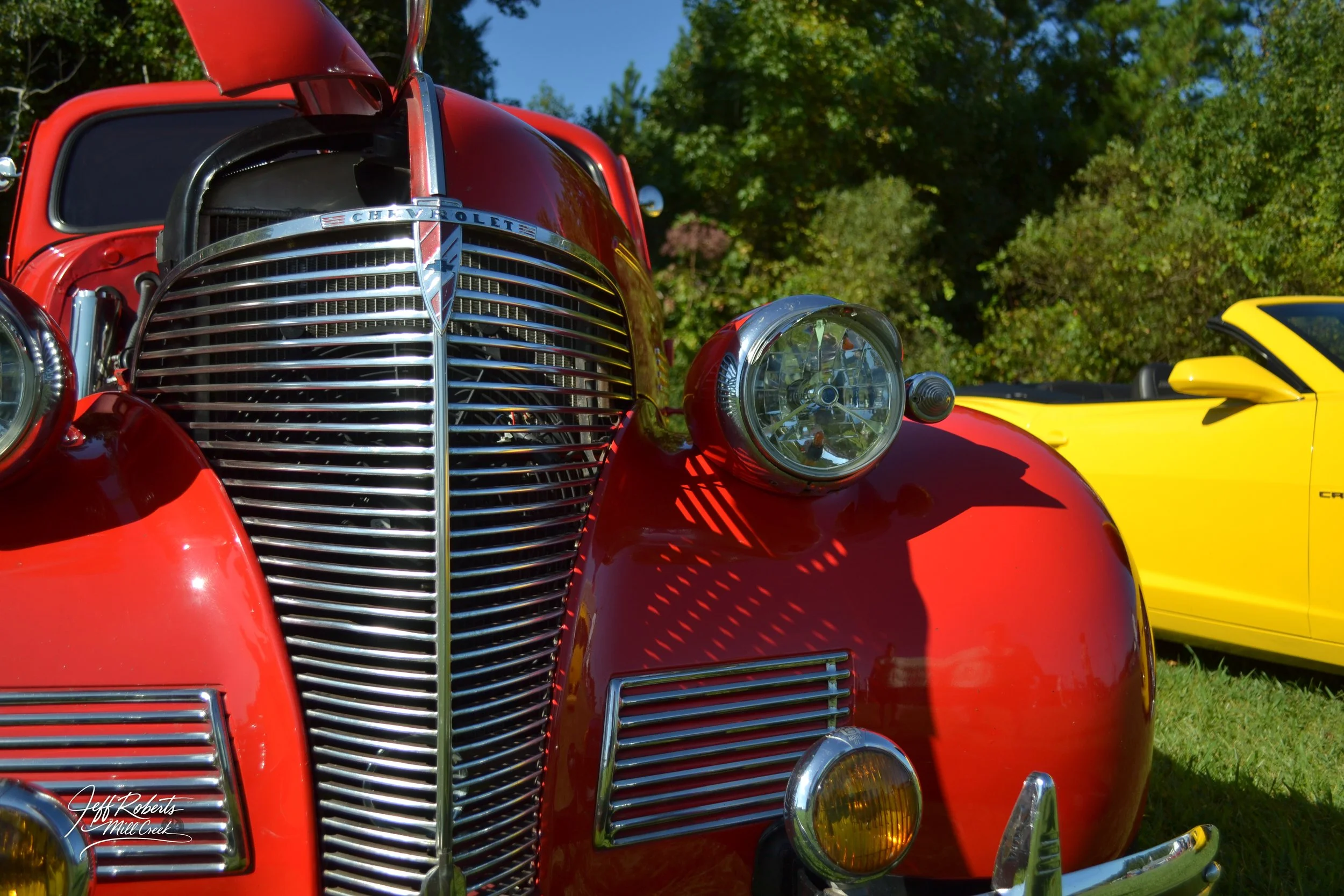 Close-up of a vintage red Chevrolet car with chrome grille and headlight, parked on grass with trees in the background, and a yellow sports car visible to the side.