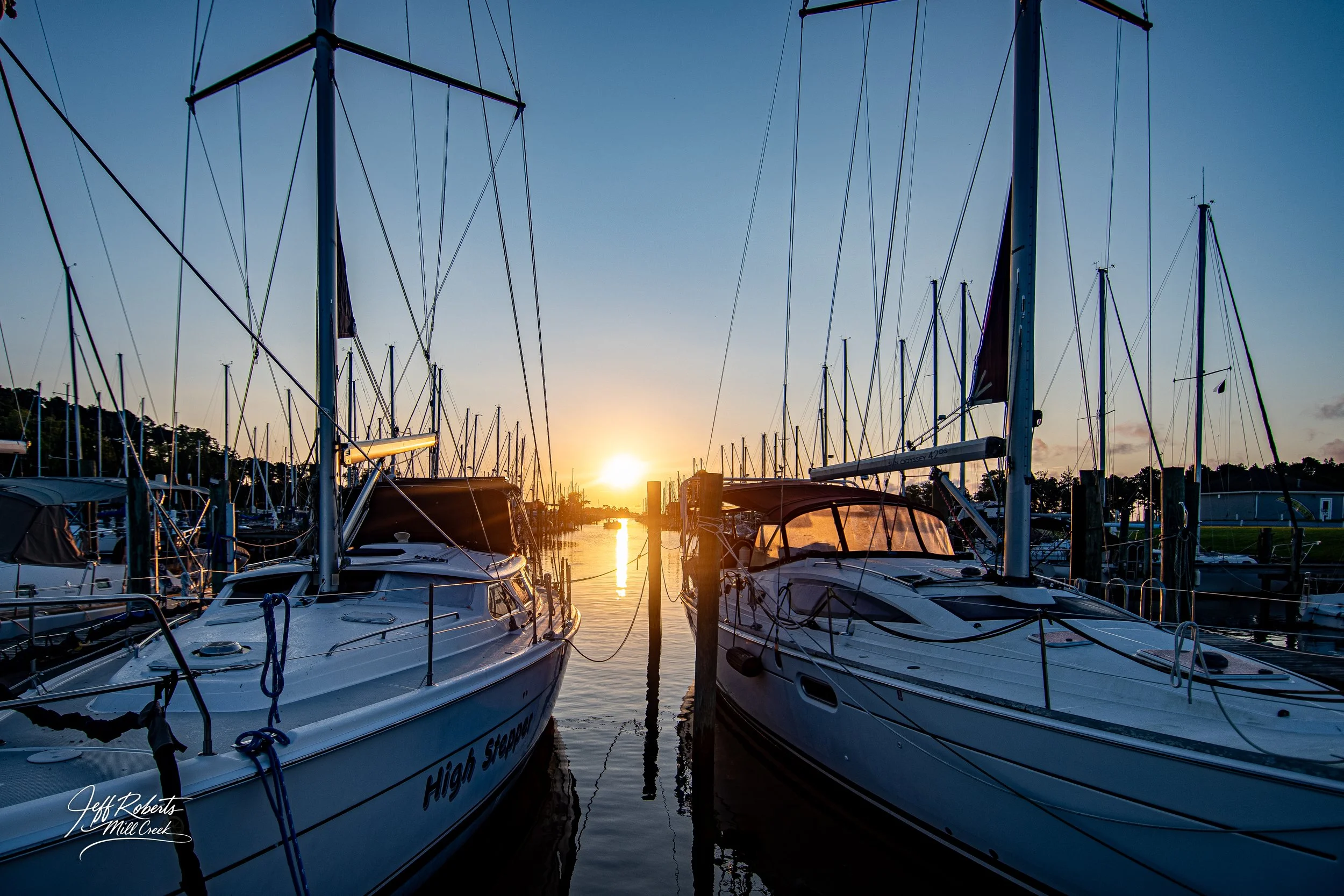 Sailboats docked at a marina during sunset with orange sky and water reflections.