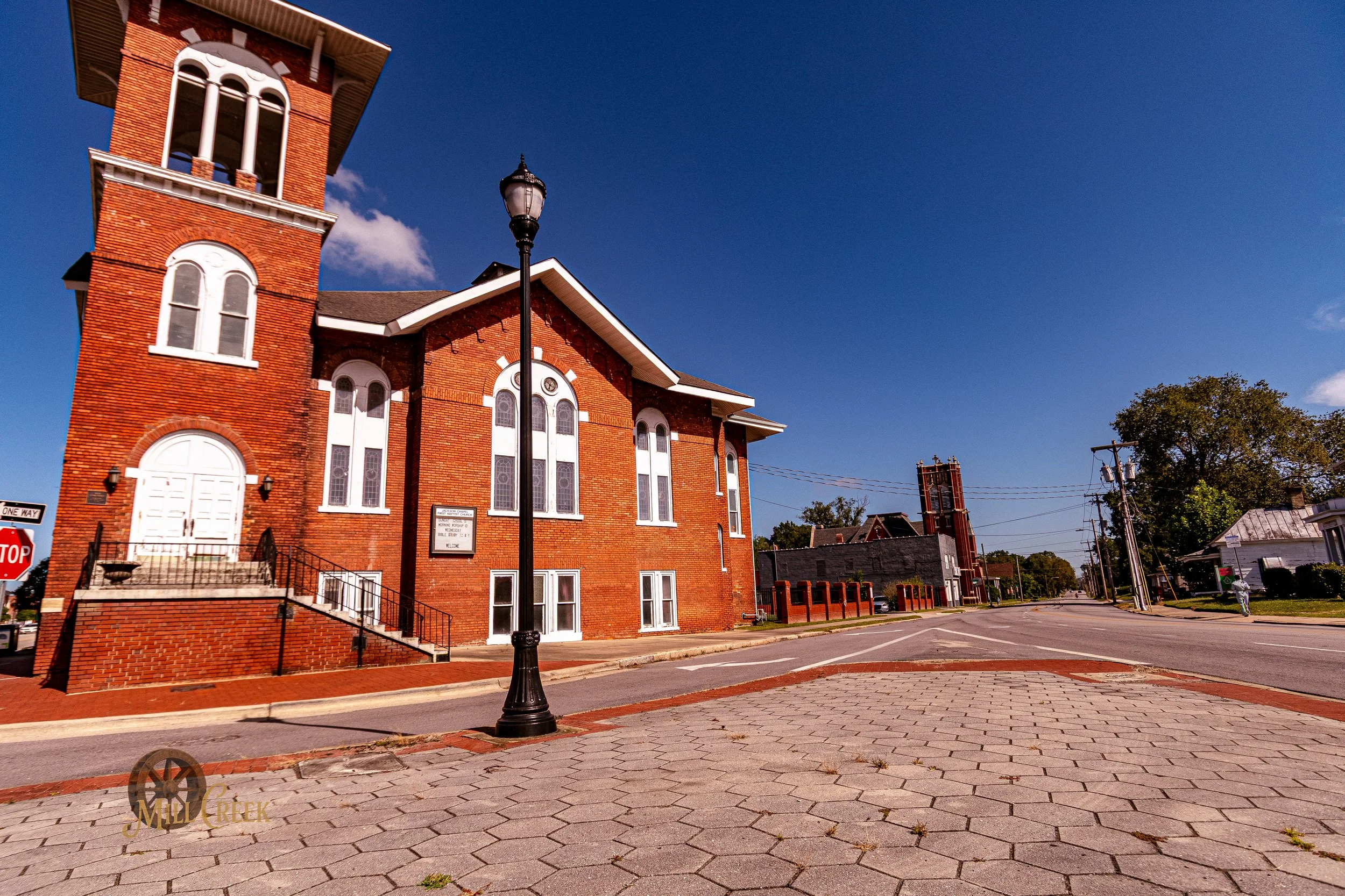 A red brick church with a tower, white arched windows, and a white door with steps and black guardrails, situated on a sunny street corner with a street lamp and a stop sign, under a clear blue sky.