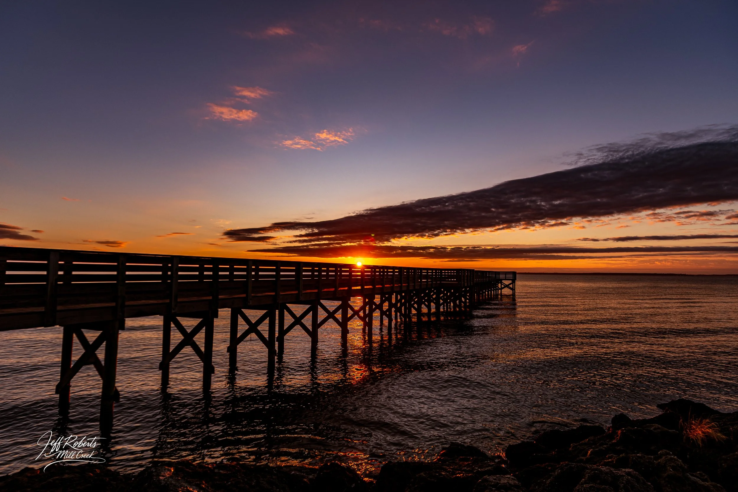 Sunset over a wooden pier extending into the water with colorful clouds in the sky.