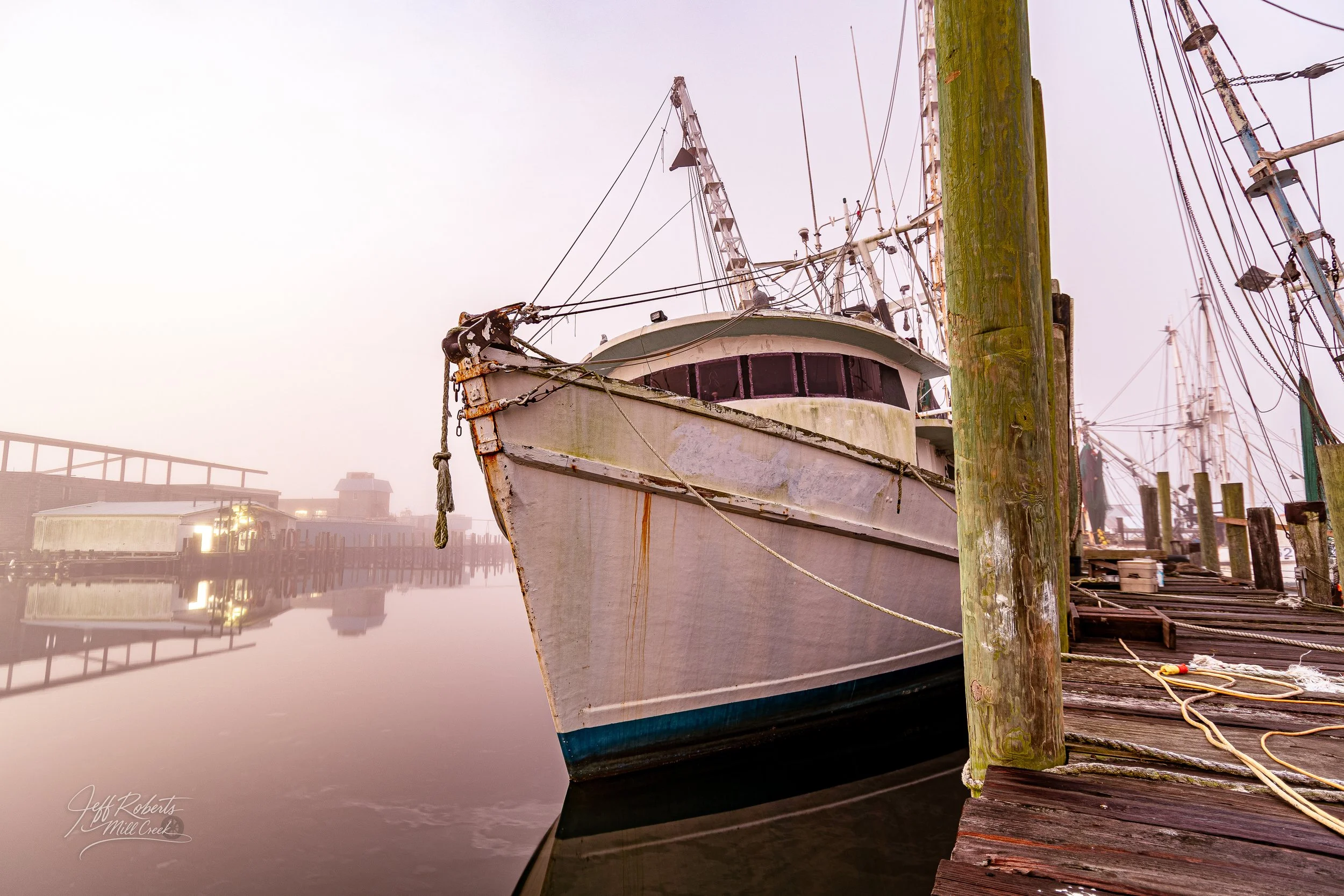 Old, weathered boat docked at a wooden pier in a foggy harbor with reflections on the water.