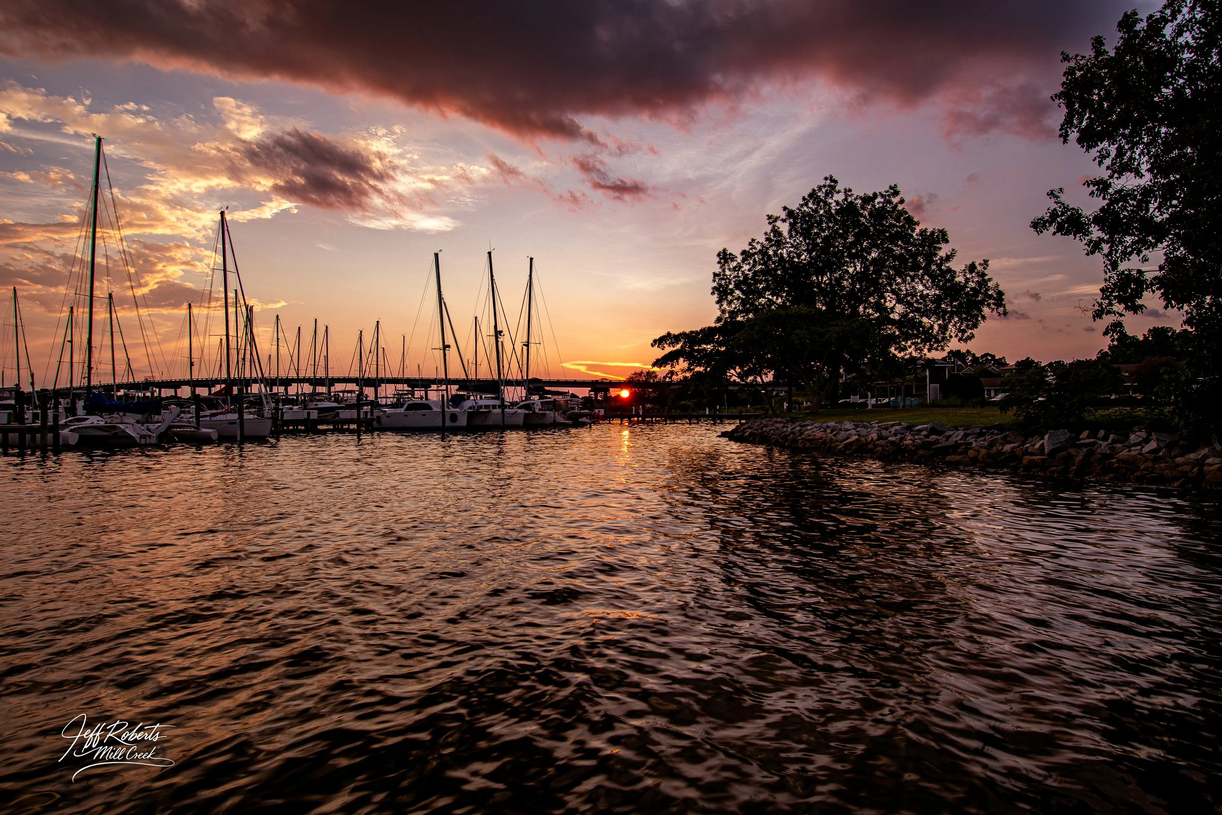 A marina at sunset with sailboats docked, calm water reflecting the sky, and trees on the shoreline, with a bridge in the background.