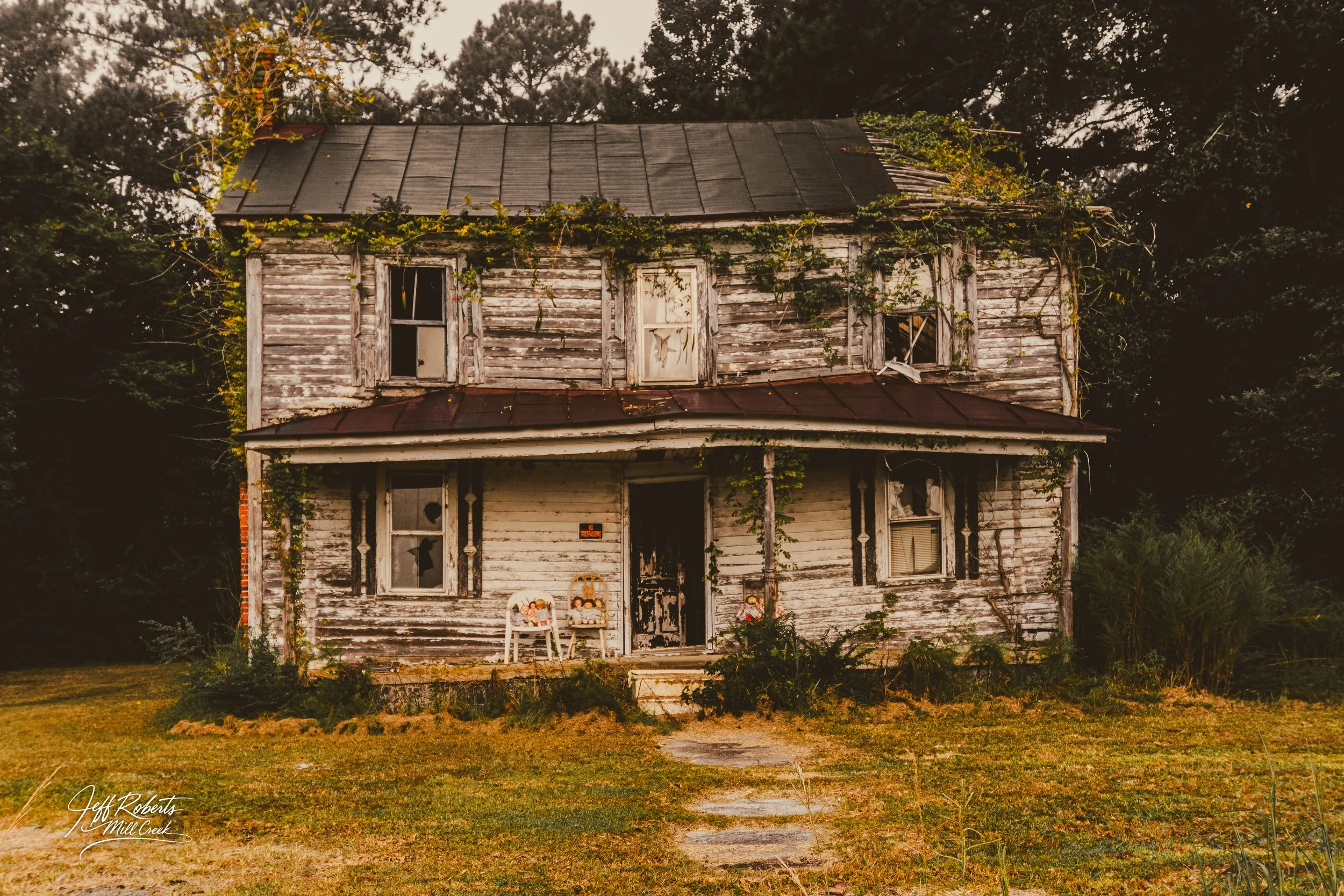 A dilapidated two-story house with peeling white paint, broken windows, and overgrown vines on the walls, surrounded by an overgrown yard.