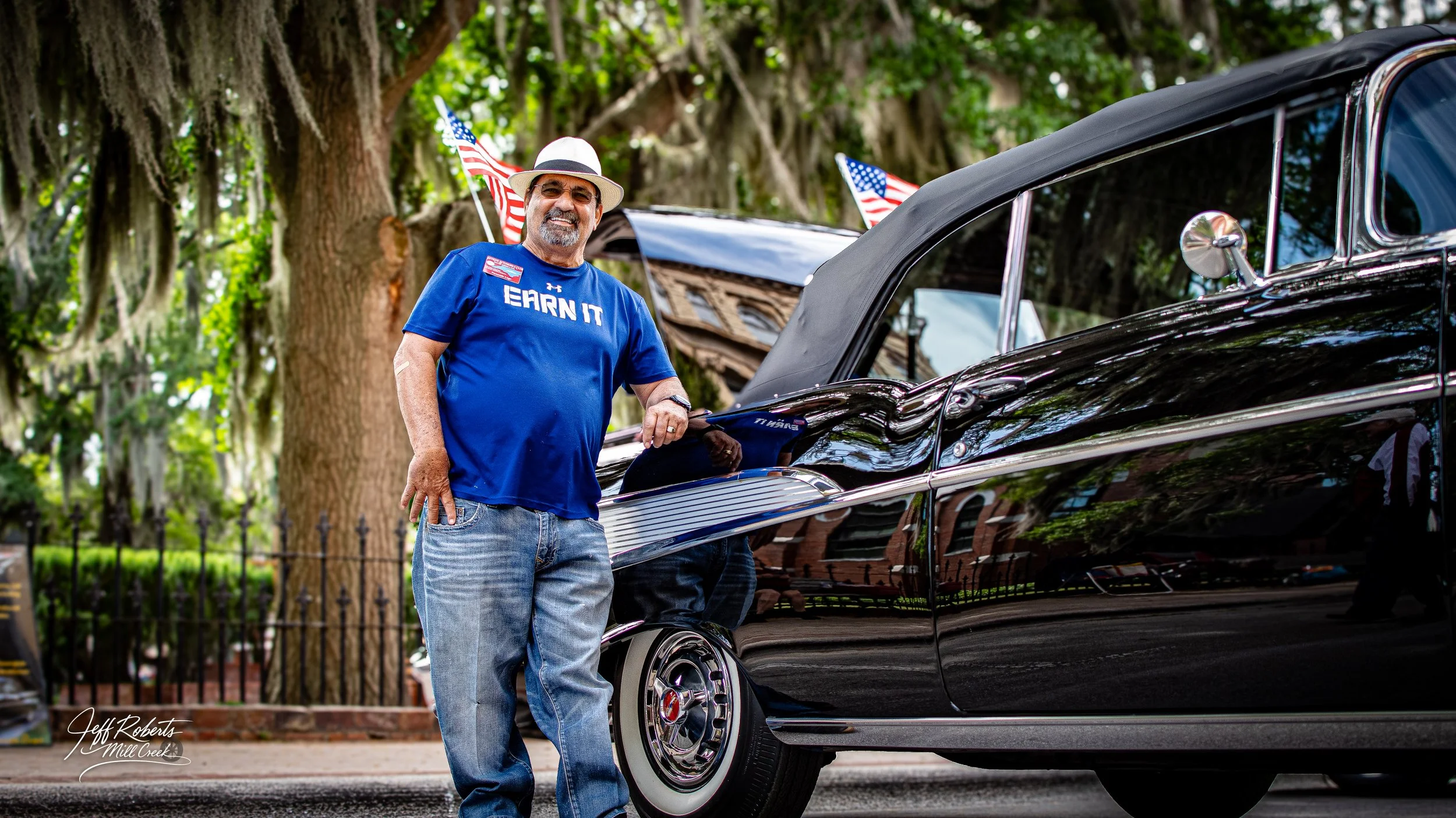 An older man in a blue t-shirt and jeans standing next to a black vintage car, smiling, with American flags in the background and trees overhead.