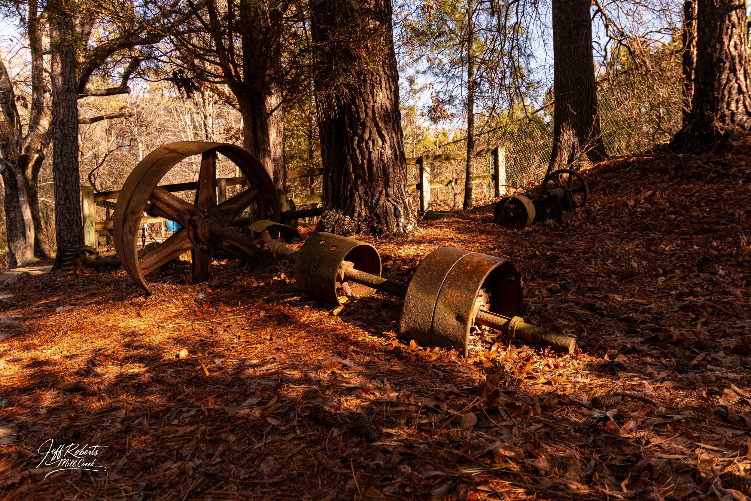 Rusty old farm equipment lying on the ground among fallen leaves in a wooded area with tall trees and a fence in the background.