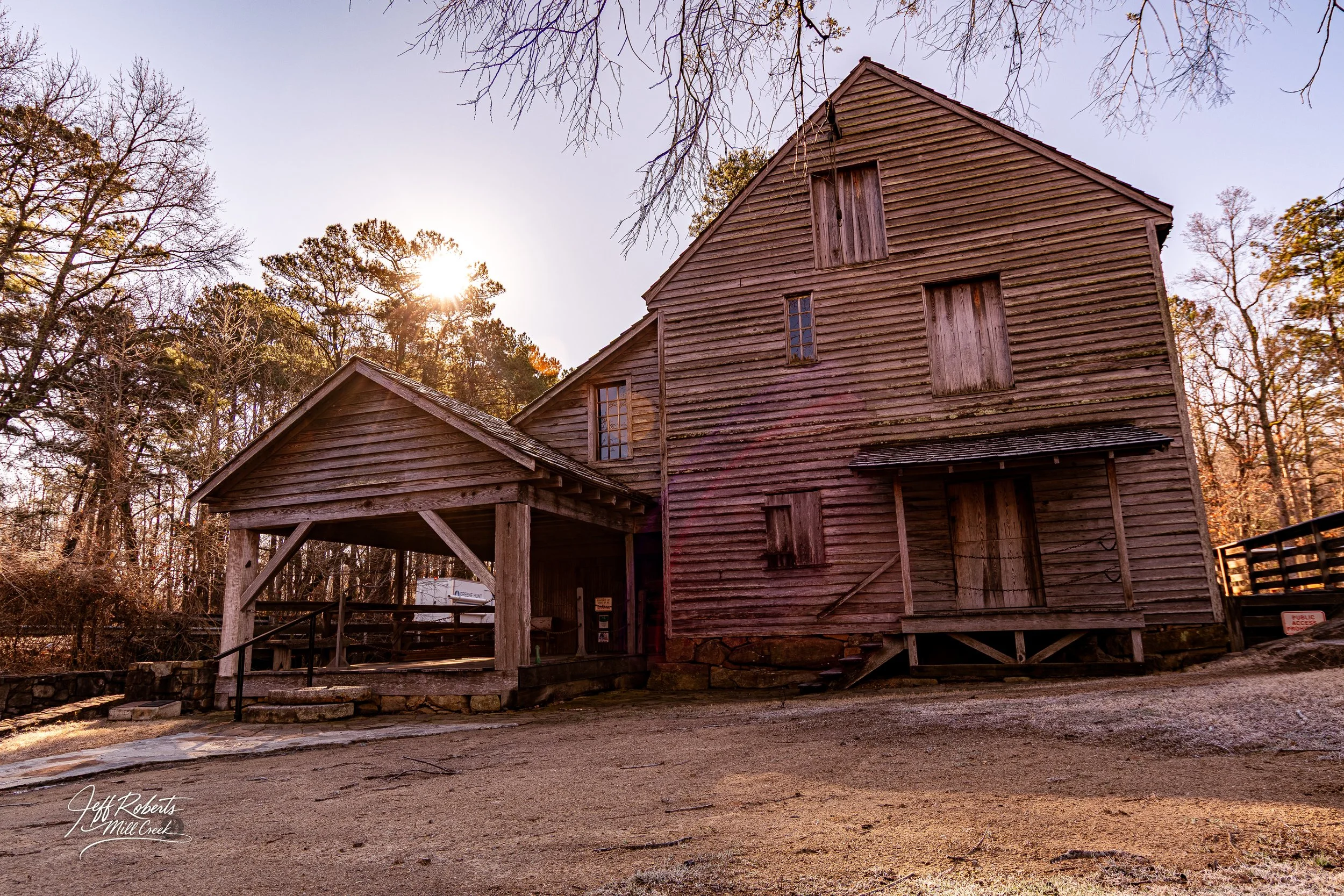 A rustic, weathered wooden mill building with boarded windows, set in a wooded area with leafless trees and sunlight peeking through, capturing a fall or winter scene.