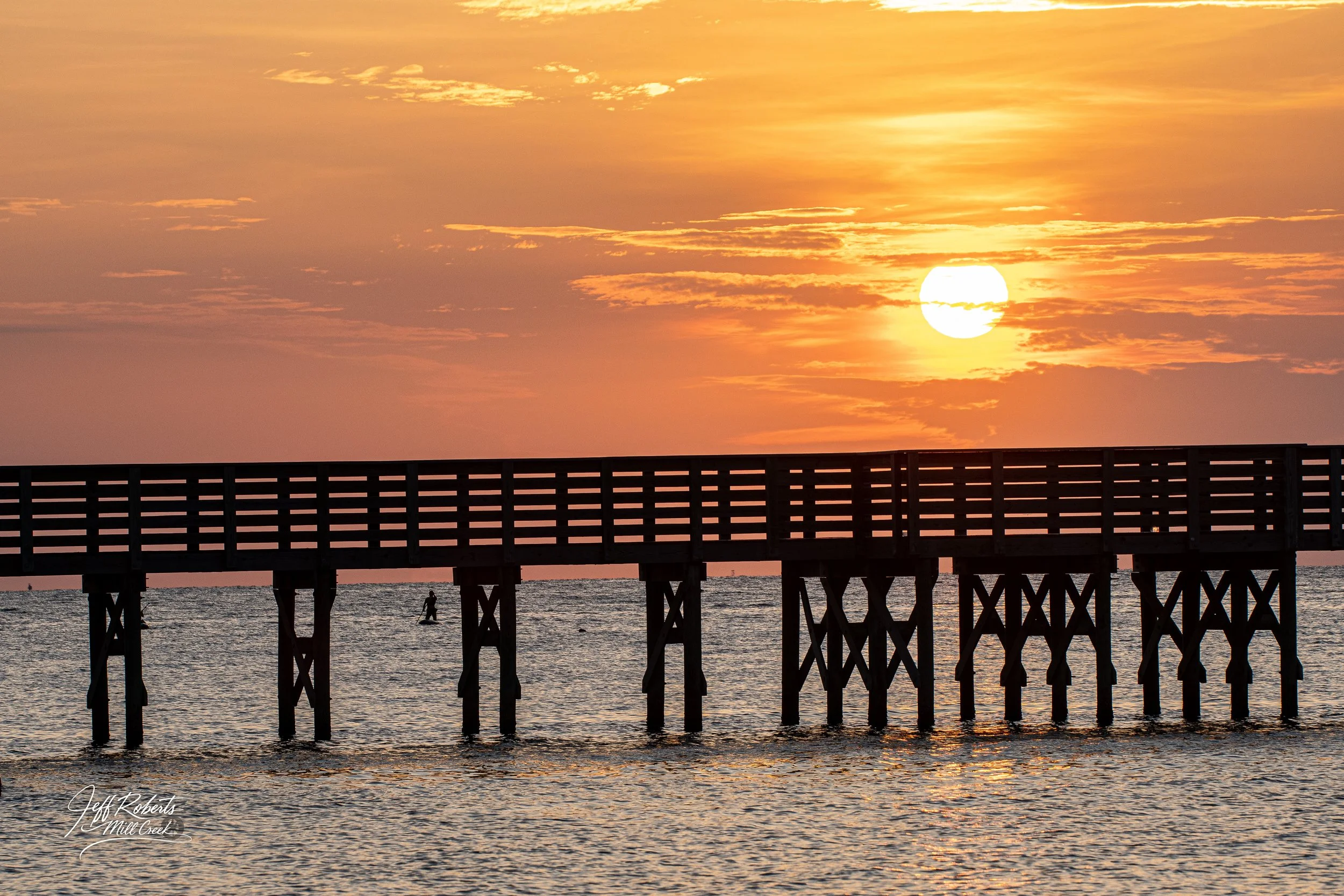 Sunset over a wooden pier extending into the water, with the sun partially covered by clouds and a person paddleboarding in the distance.