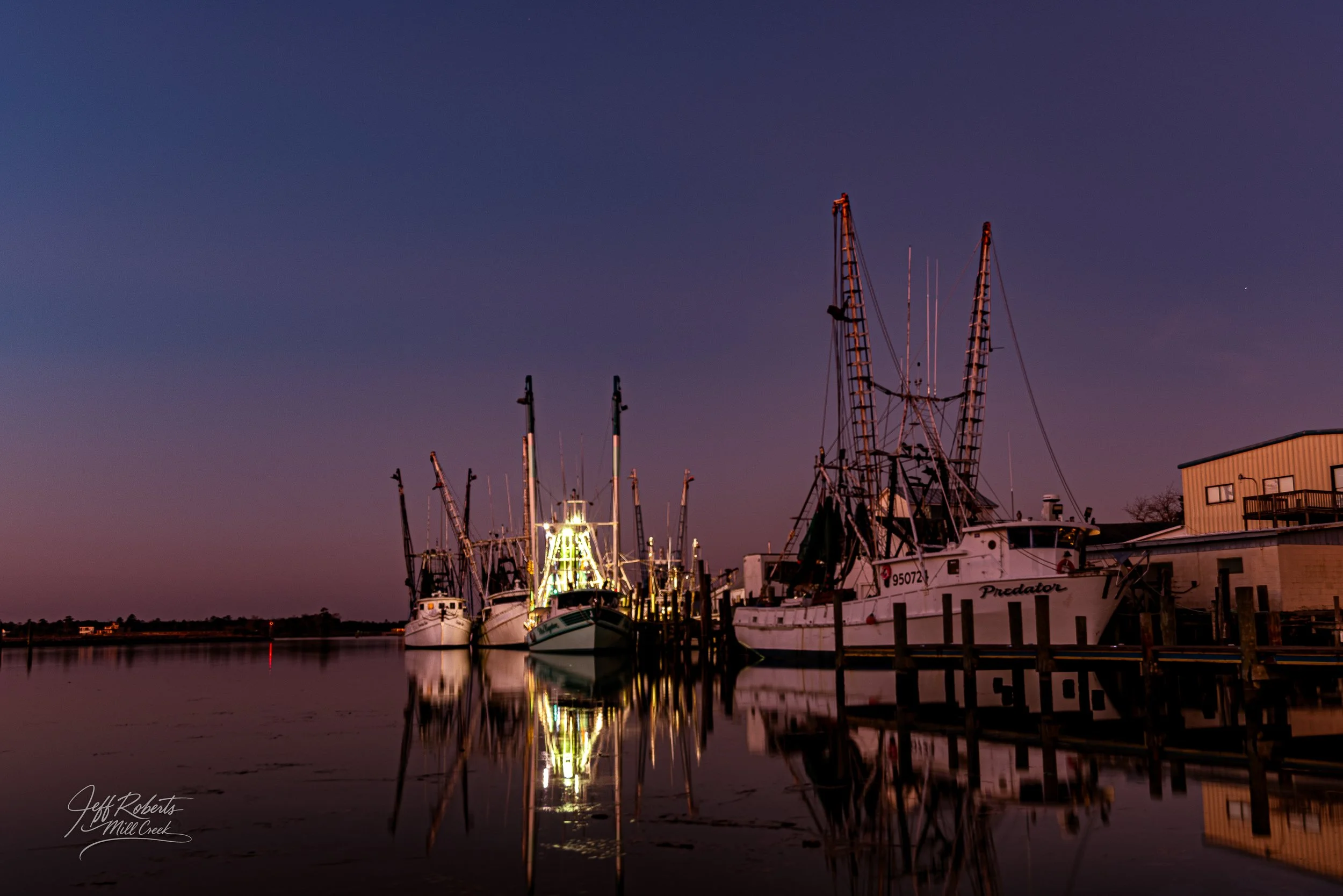 Nighttime scene of fishing boats docked at a pier with their reflections in calm water, illuminated by lights, under a darkening sky.