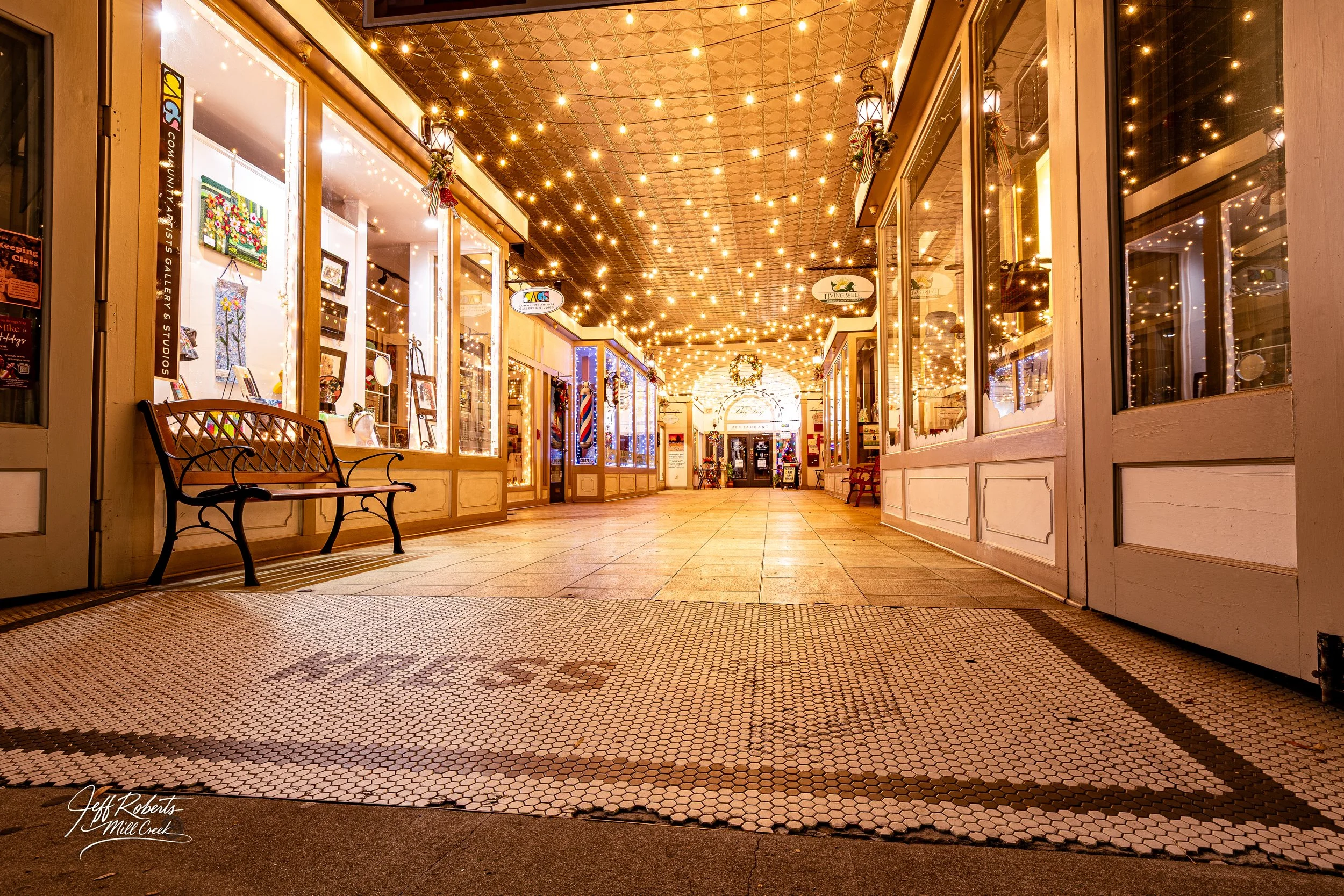 Empty indoor shopping arcade decorated with warm string lights, featuring small boutique shops, benches, and a tiled floor with decorative patterns.