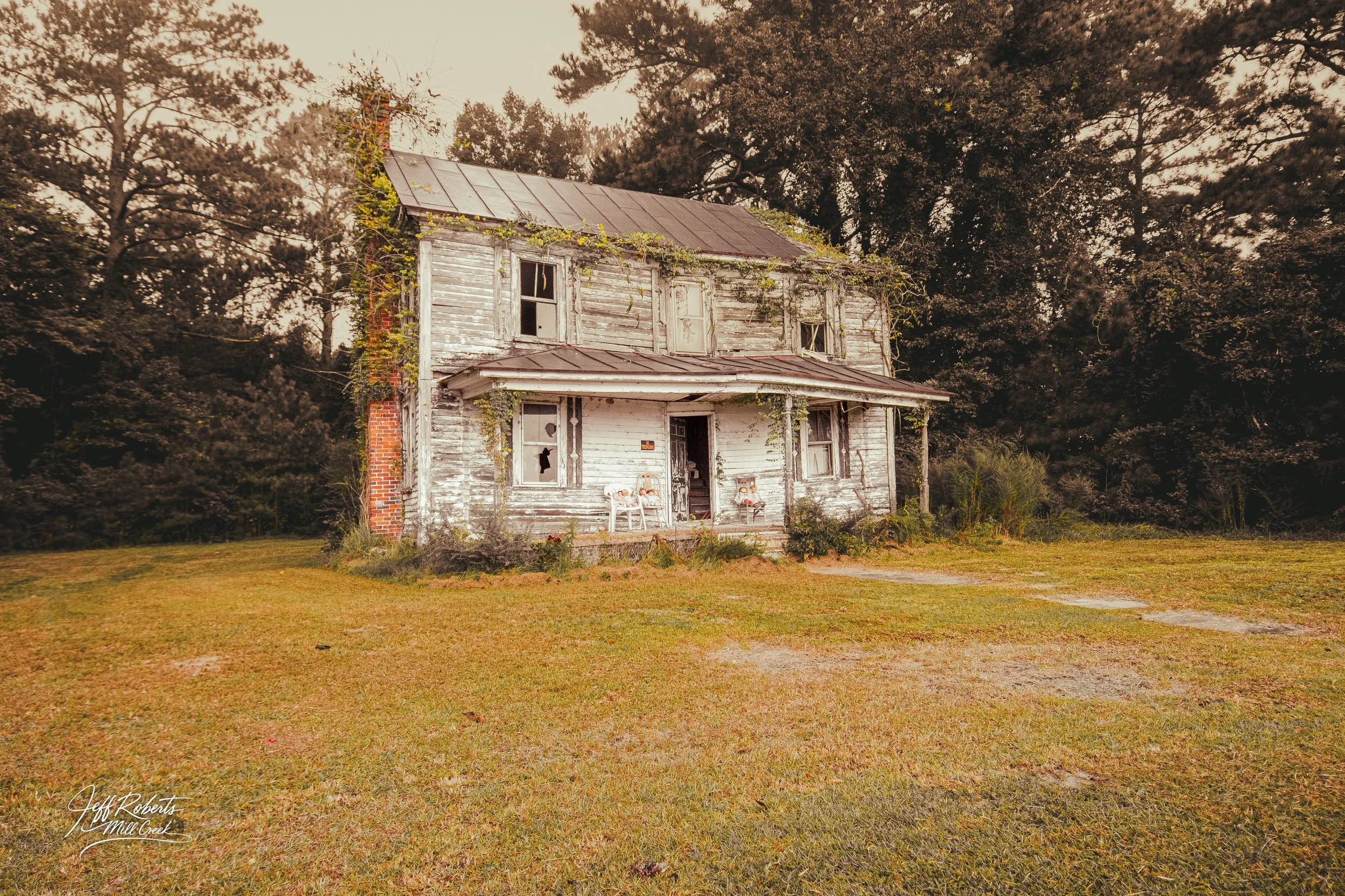 An old, abandoned two-story wooden house with peeling paint, broken windows, and a rusty roof, surrounded by overgrown vegetation and trees in the background.